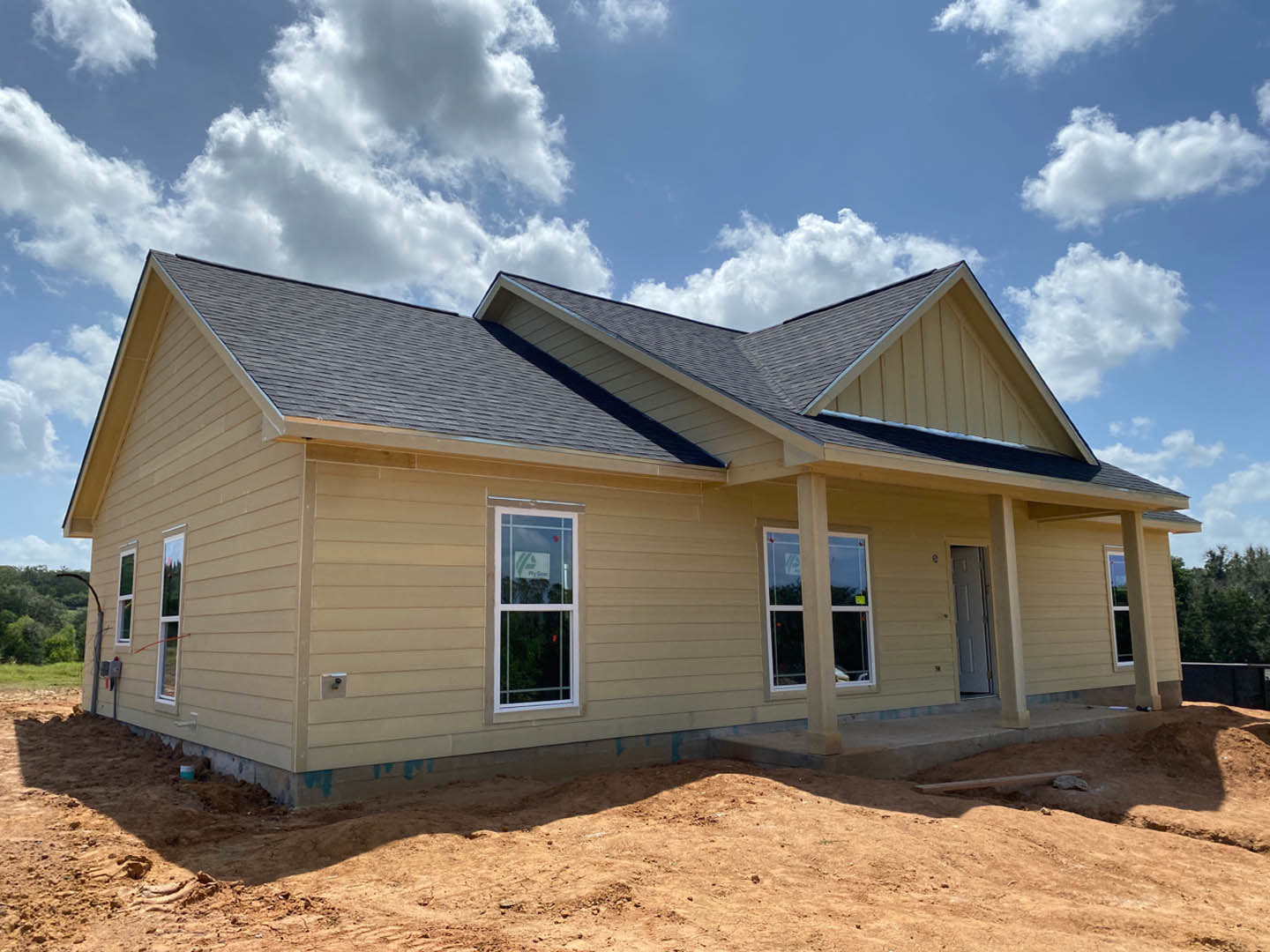 Framed custom home under construction with exposed wood, dirt lot in foreground, and partly cloudy blue sky overhead