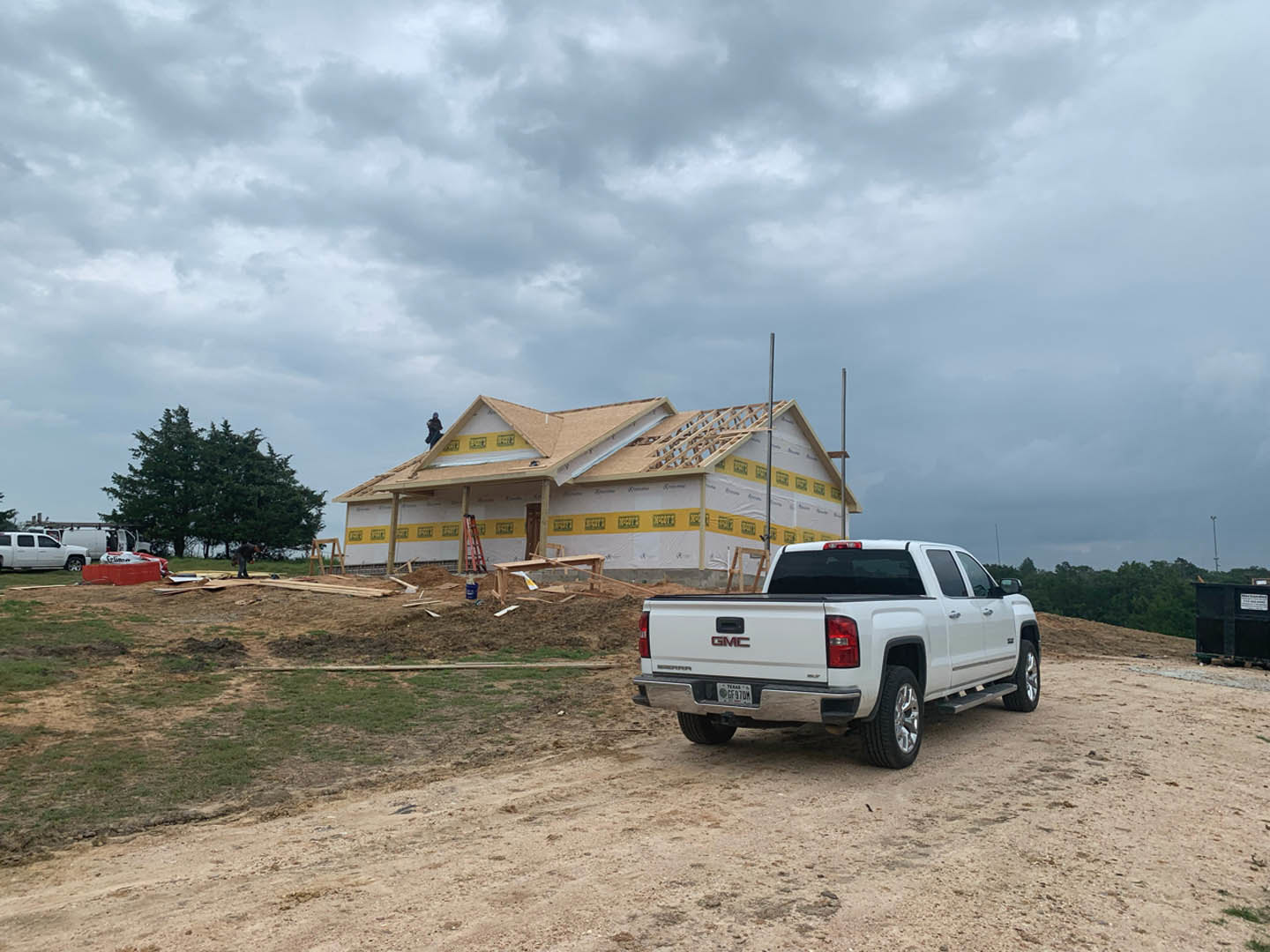 White pickup truck parked in front of partially built house with exposed framing, ladder leaning against exterior, worker on roof, grassy yard and trees in background
