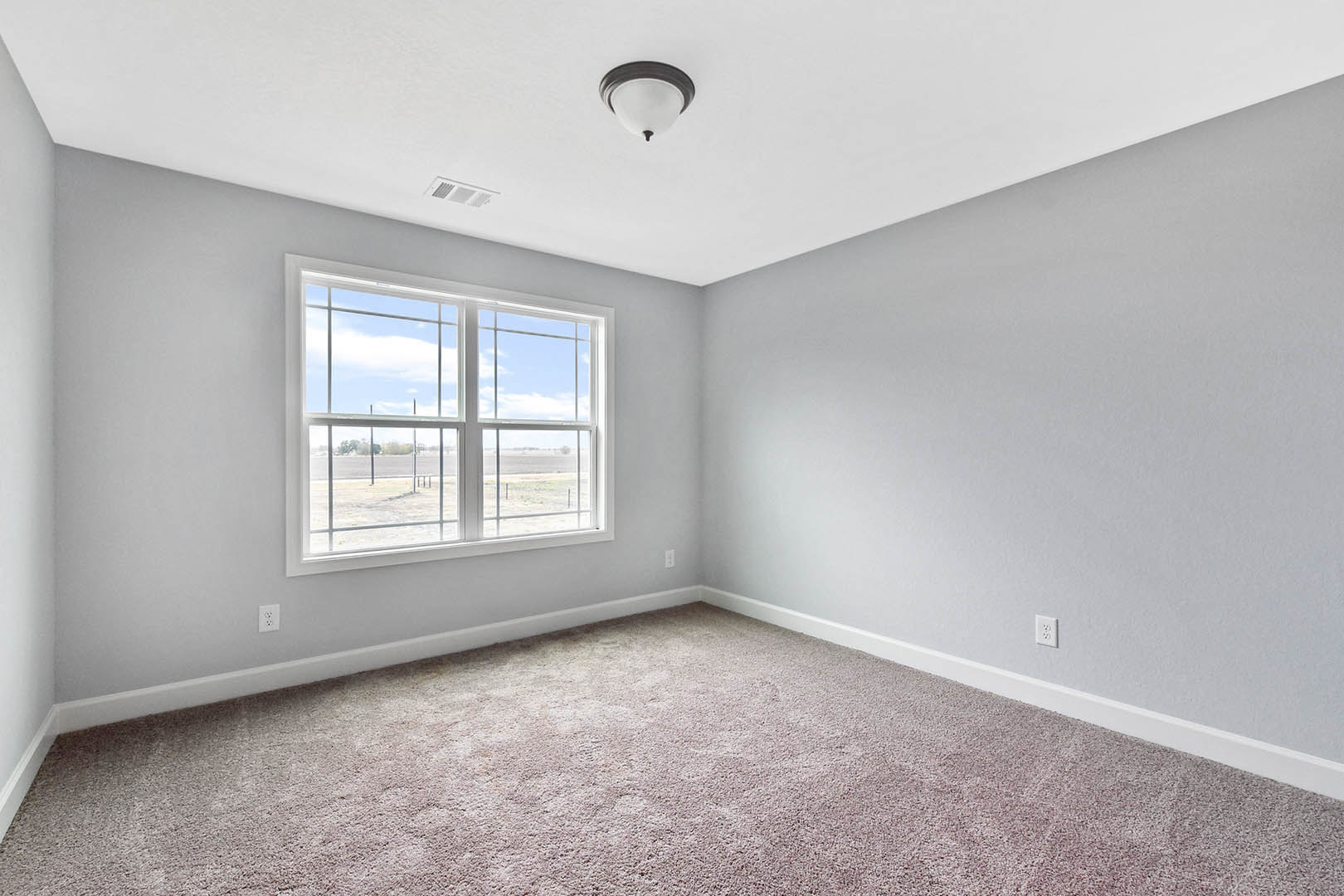 Neutral-toned carpeted room with white walls, large window overlooking open field and sky, ceiling-mounted light fixture, simple molding along ceiling and baseboards