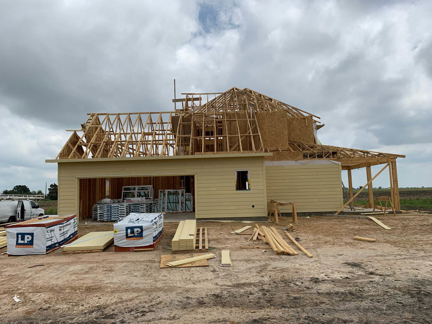 Partially built house with exposed framing, white tarp covering pallets, large white and blue tent, open white van parked nearby, cloudy sky overhead