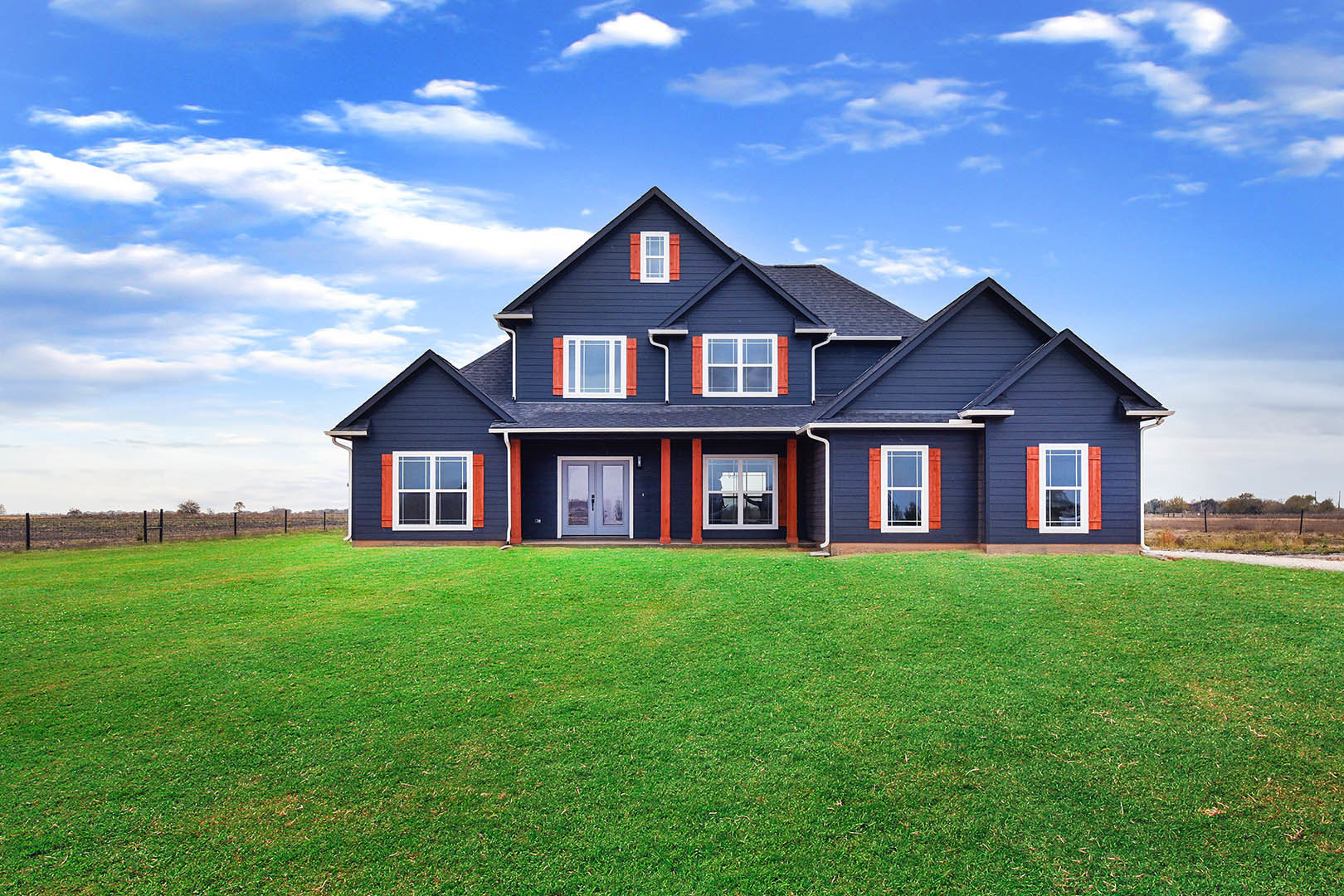 Blue house with red trim and white-framed windows, double front doors, green lawn, cloudy sky