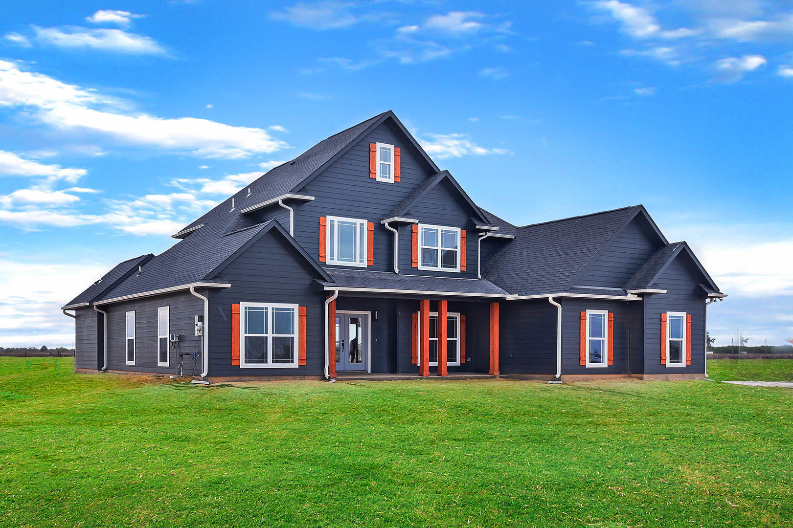 Two-story farmhouse with blue roof, orange and red window shutters, red pillars, glass door, and green lawn under partly cloudy sky