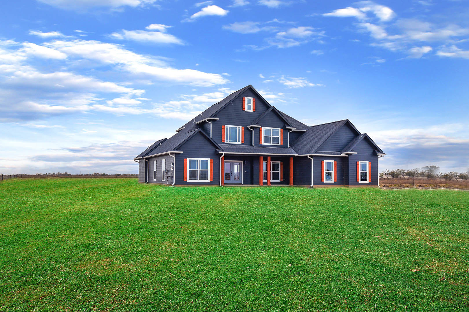 Blue farmhouse with red trim, expansive green lawn, white-framed windows, blue shutters, and partly cloudy sky