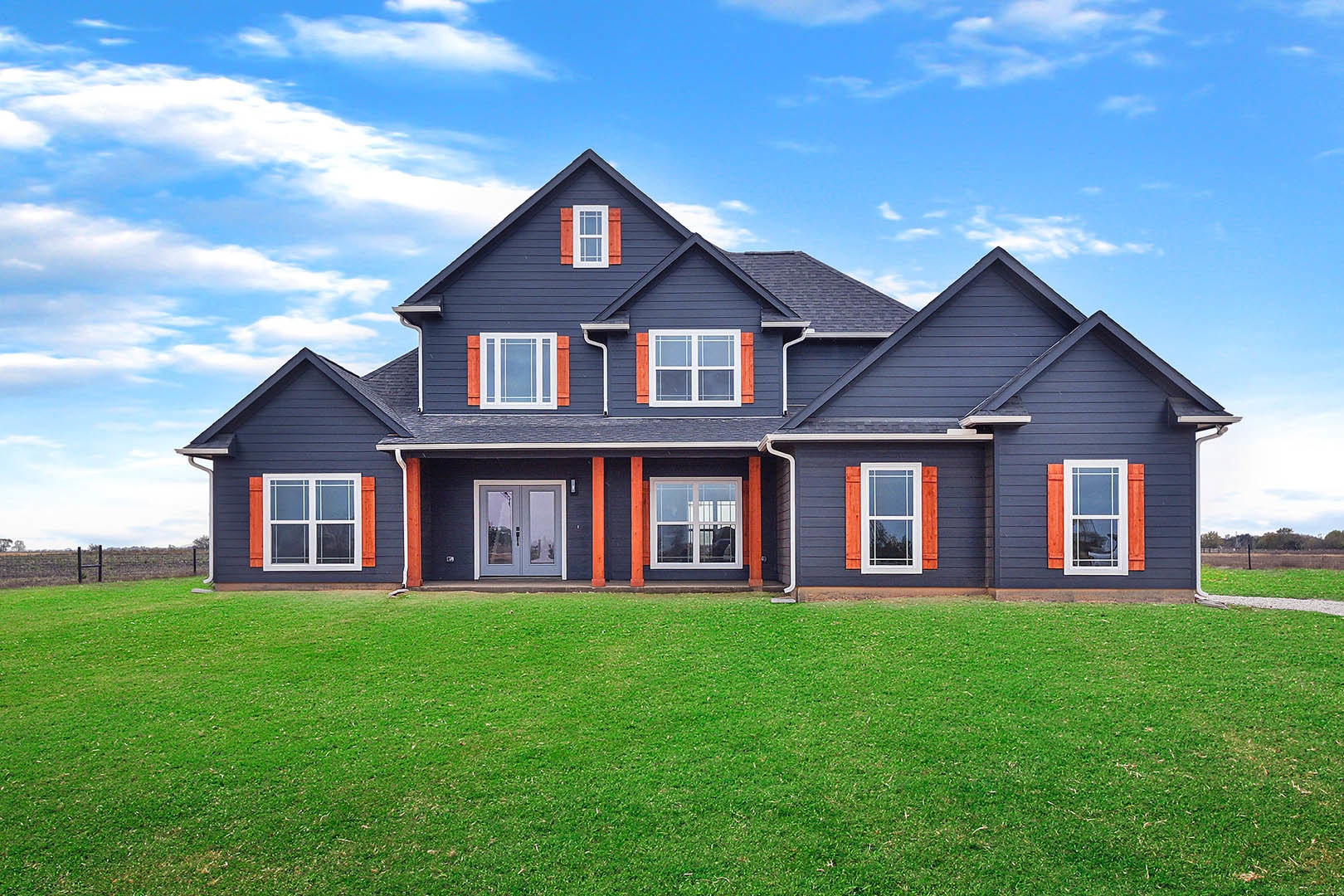 Two-story house with blue roof, white-framed windows, double front doors, orange trim, and green lawn under partly cloudy sky