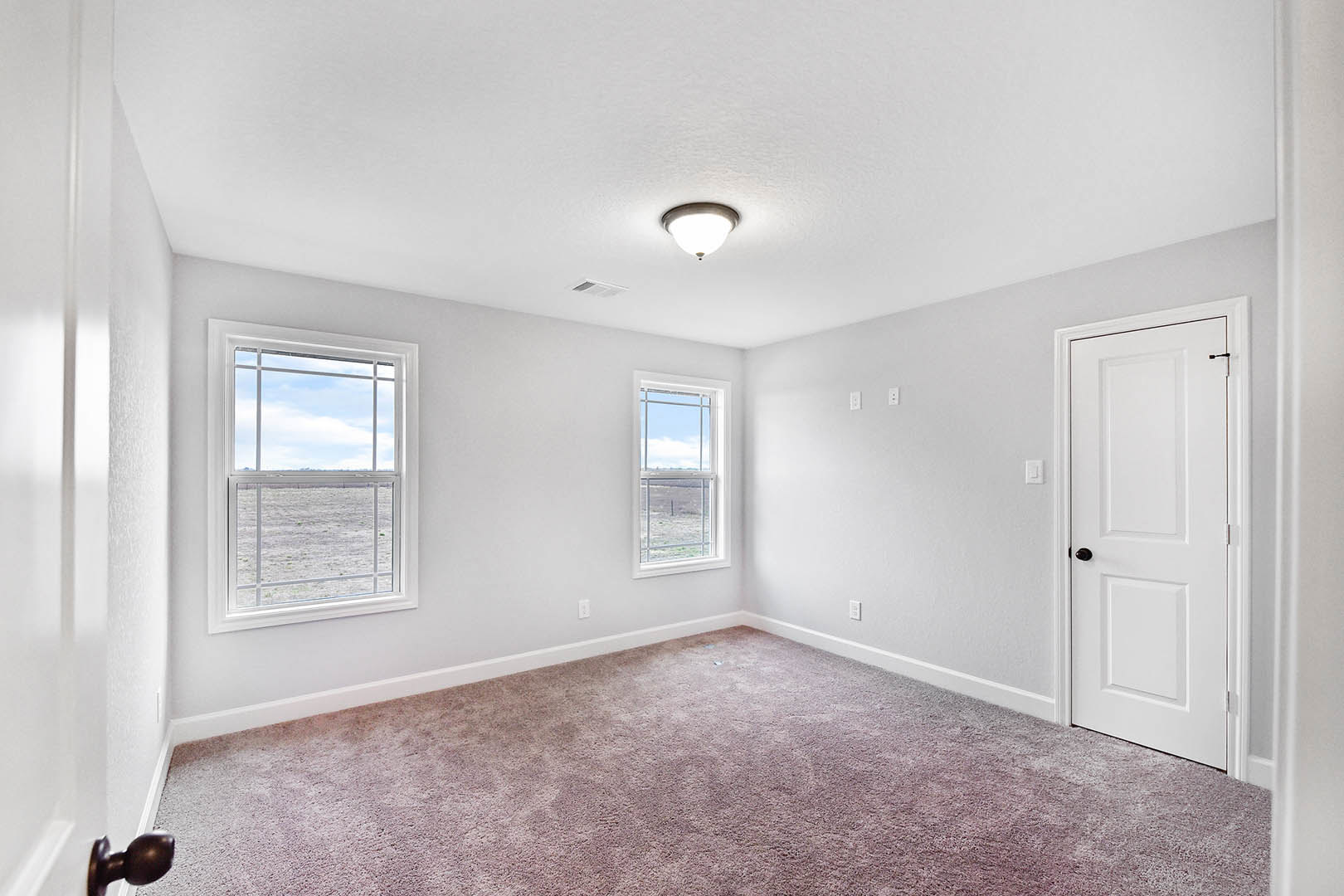 Bright room with two large windows overlooking a grassy field, white paneled door with black handle, neutral carpet flooring, simple ceiling light fixture, smooth white plaster