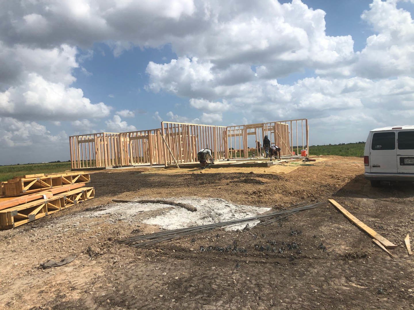 Framed house under construction with exposed wooden beams, white van parked nearby, worker on site, cloudy sky overhead, scattered lumber on dirt ground