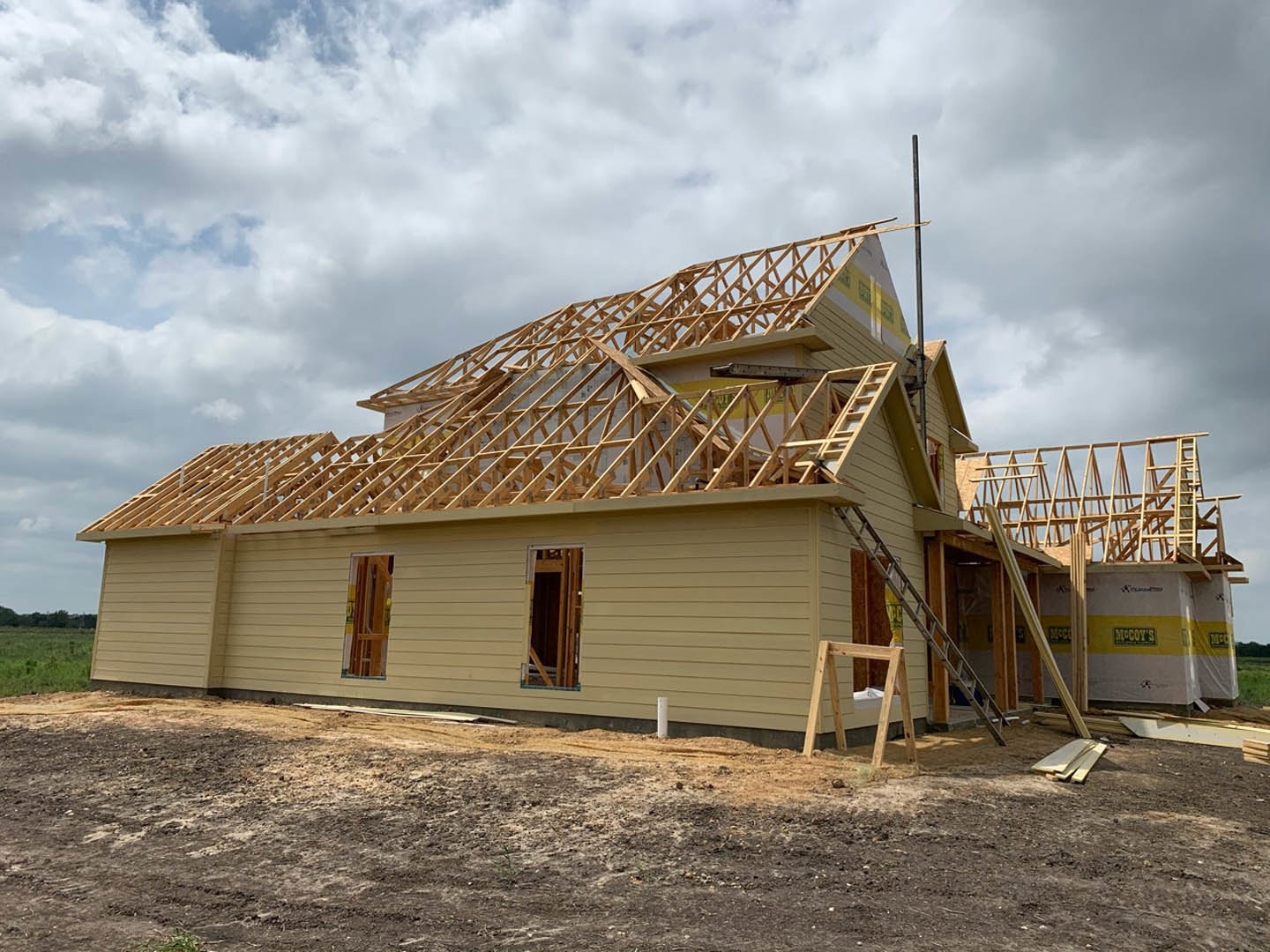 Wood-framed house under construction with exposed beams, ladder leaning against unfinished wall, dirt patch and pile of wood planks in foreground, cloudy sky overhead
