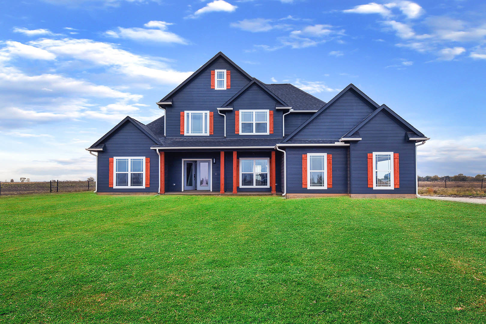 Blue farmhouse with red shutters, white-framed windows, and a green lawn under a partly cloudy sky