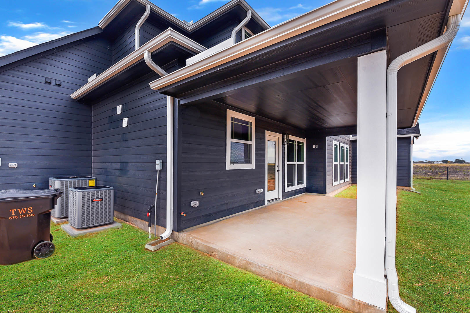 Two-story home with white siding, covered front porch, green lawn, black waste container near entrance, multiple windows, and visible air conditioning units along the side
