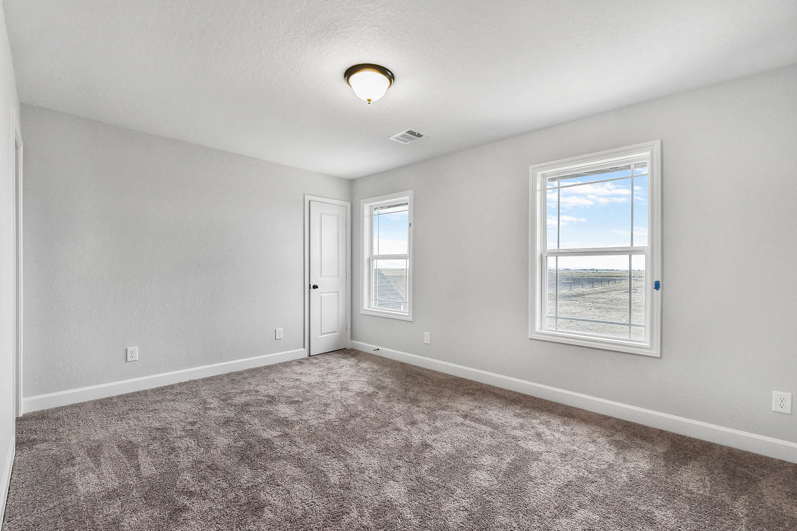 Neutral-toned carpeted room with white walls, large window overlooking a grassy field, white door with black hardware, and ceiling light fixture