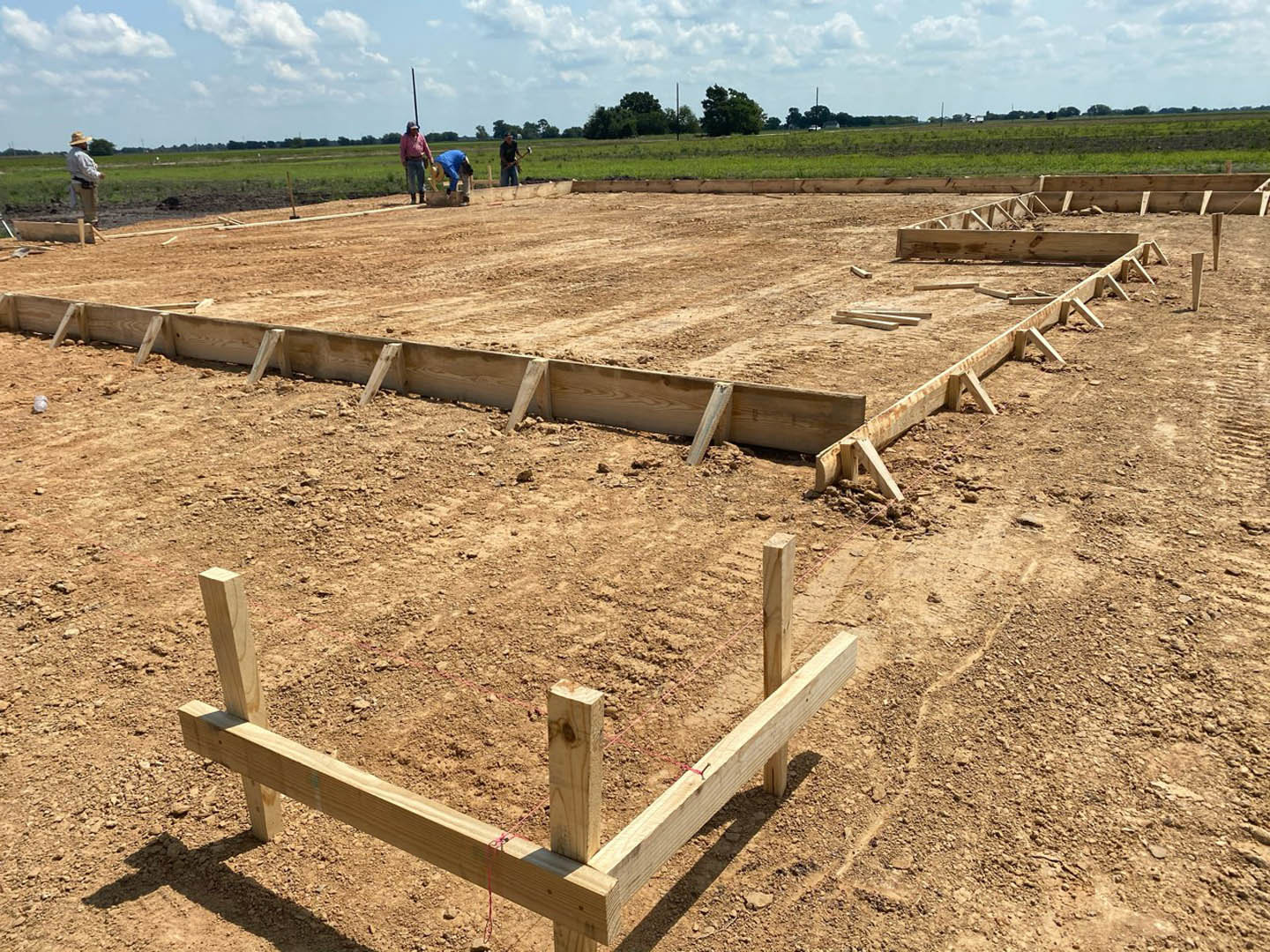 Wooden framing and foundation posts set on dirt, red string marking boundaries, workers assembling structure, cloudy sky and tree line in background