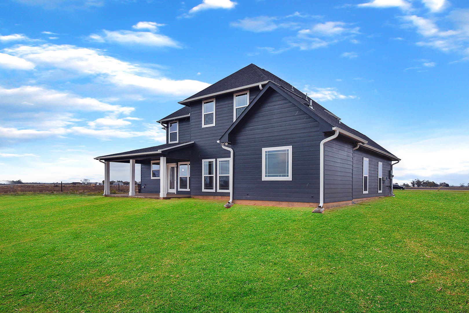Two-story grey house with blue roof, white-framed windows, and manicured green lawn under a partly cloudy sky