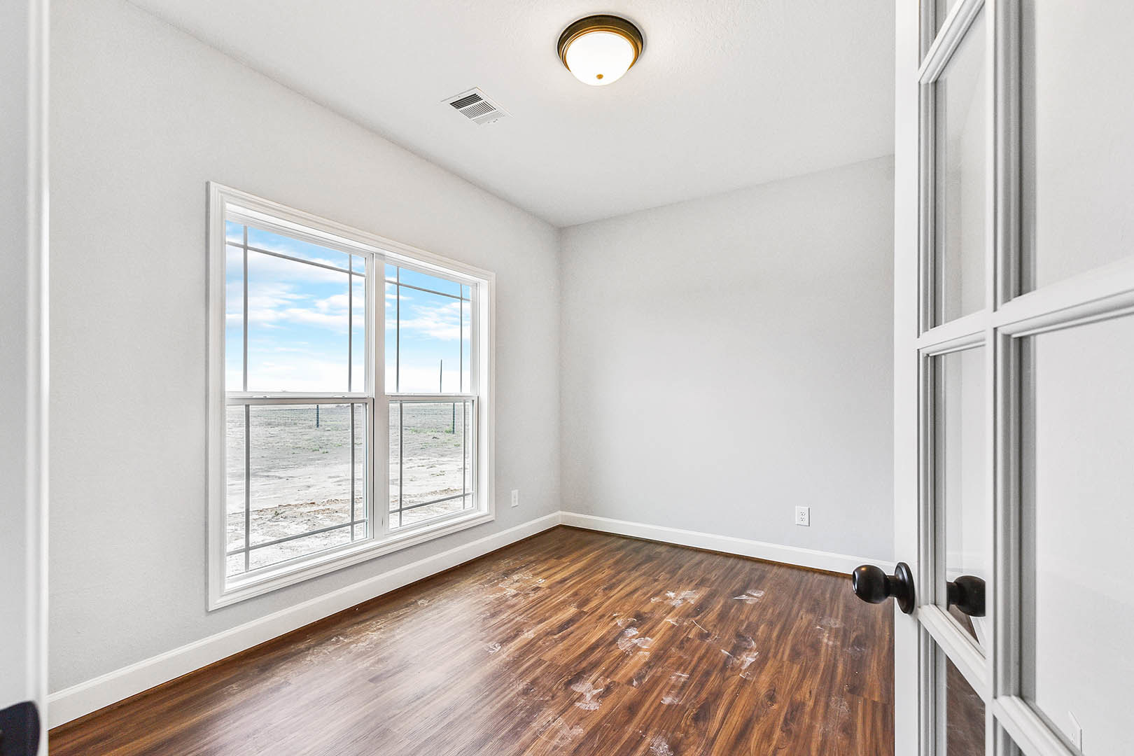 Sunlit room with wide plank wood flooring, large window overlooking desert landscape, white painted walls, ceiling vent, modern black door knob, and contemporary light fixture.