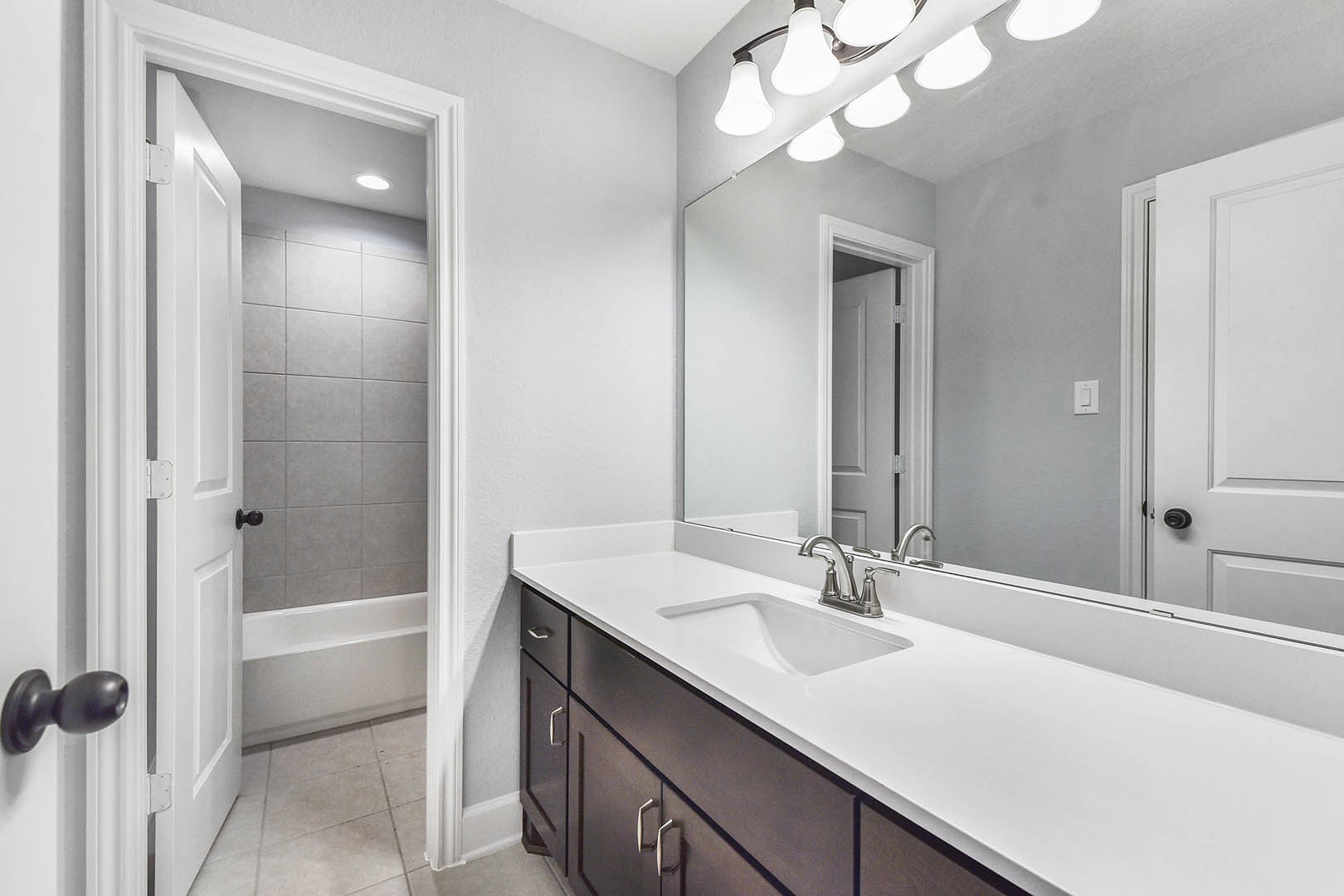 White bathroom with tile walls, modern sink and chrome faucet, large mirror above countertop, open door with brushed metal doorknob, ceiling light fixture visible