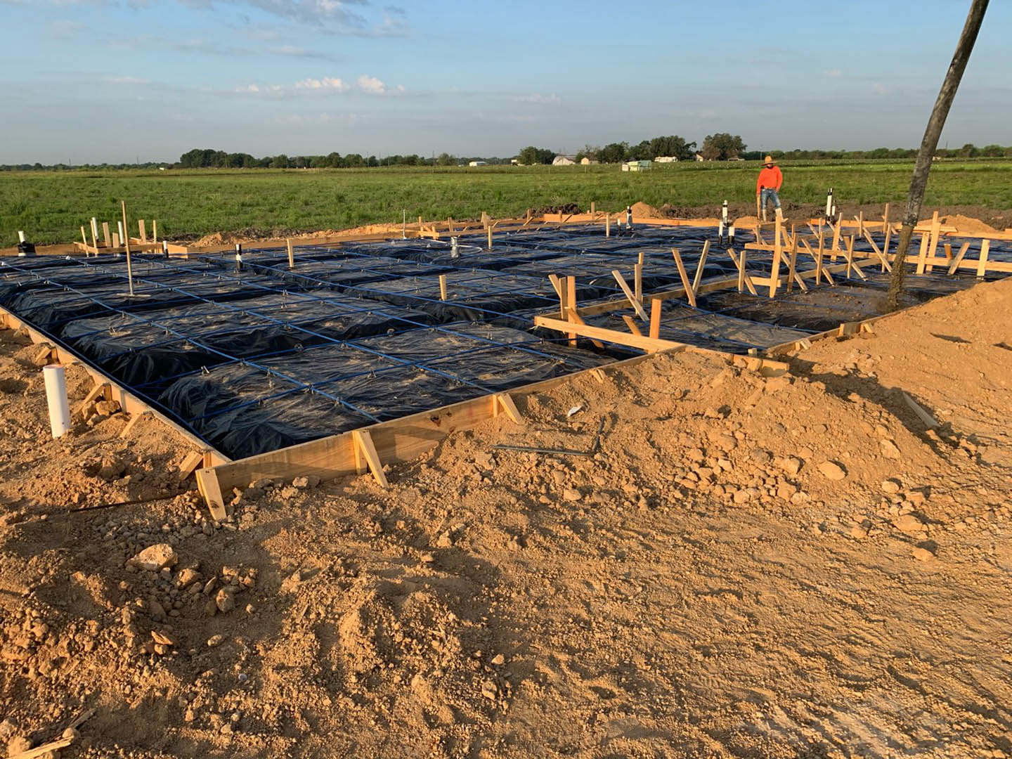 Man standing in front of a residential construction site with exposed wooden beams, black tarp, and blue plastic sheeting under a partly cloudy sky, grassy ground and tree trunk