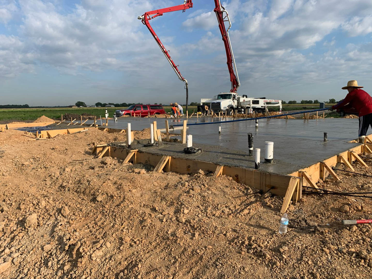 Framed custom home under construction with exposed wood beams, concrete foundation, crane lifting materials, workers in hard hats, trucks parked on dirt lot, cloudy sky overhead