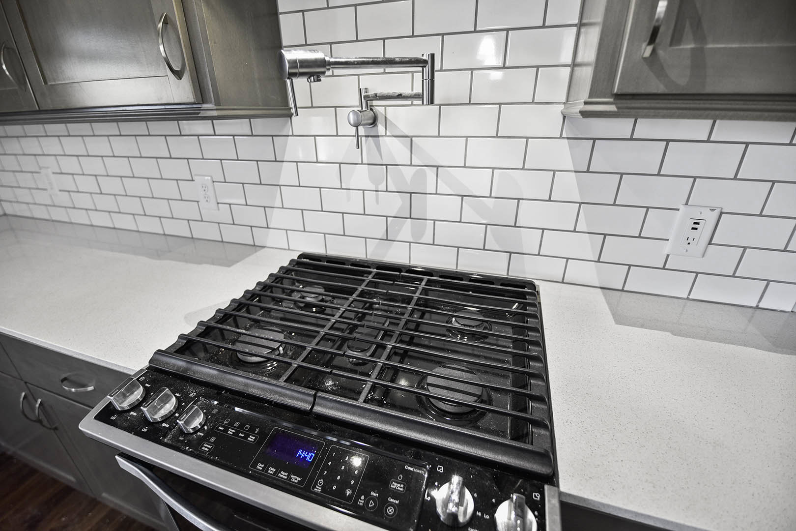 Gas cooktop with stainless steel knobs set in a tiled backsplash, modern pendant light fixture overhead, white outlet with USB ports on the wall, black oven below countertop.