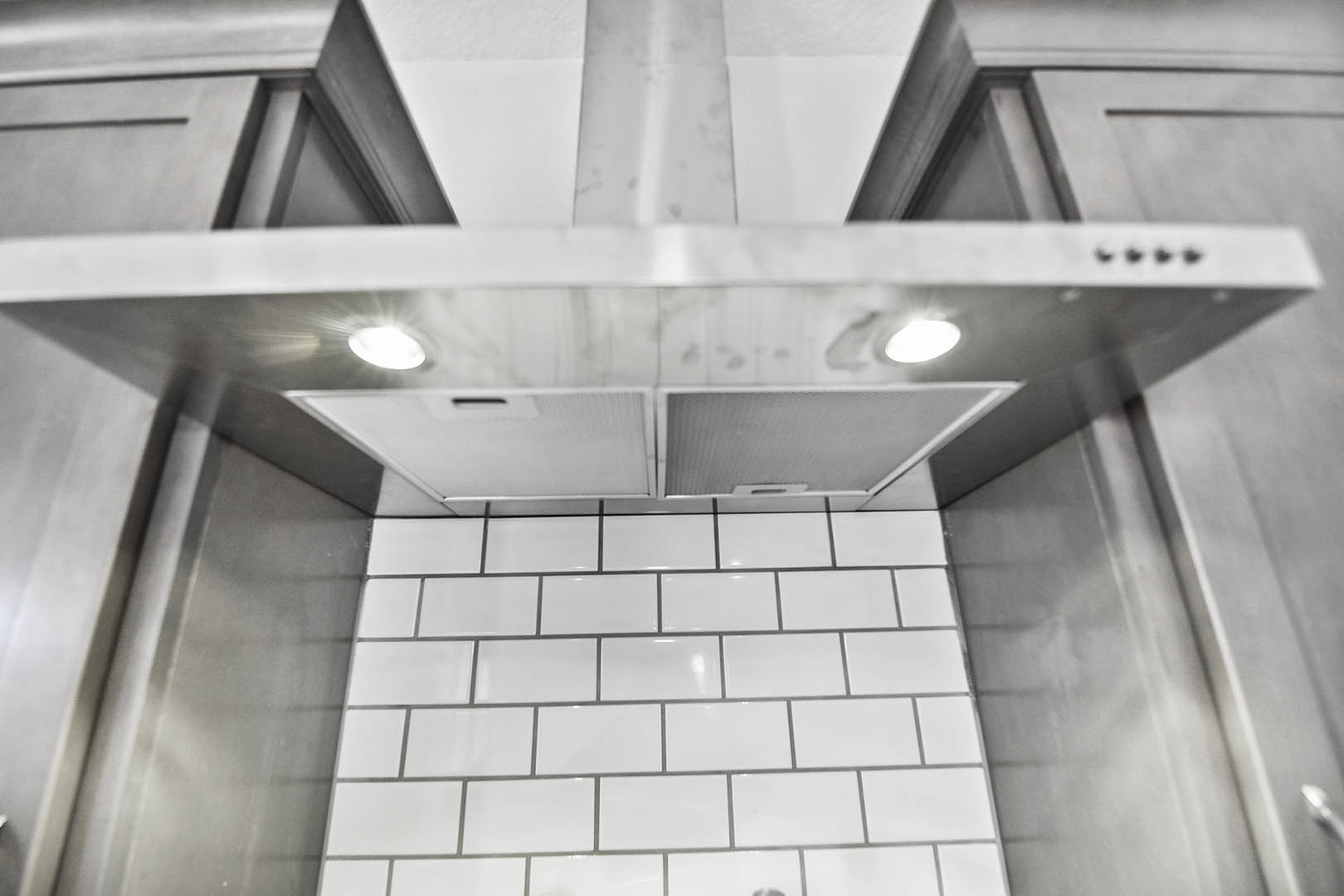 Stainless steel range hood mounted above a white tile backsplash with black grout, silver trim, and handle, under a white ceiling in a modern kitchen.