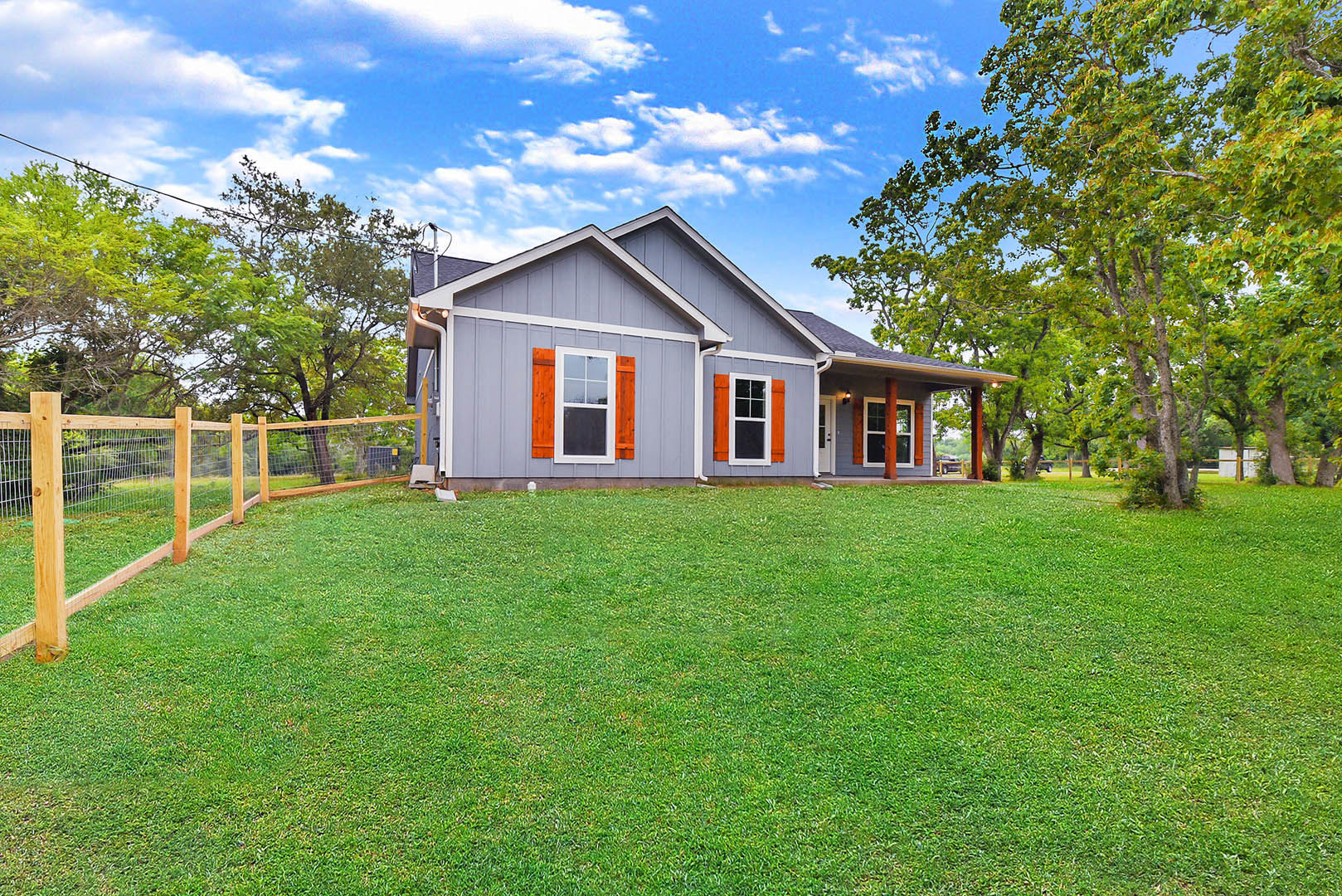 White farmhouse with orange trim, surrounded by mature trees and a grassy lawn, white fence running along a gentle hill, large window with white frame, white front door visible.