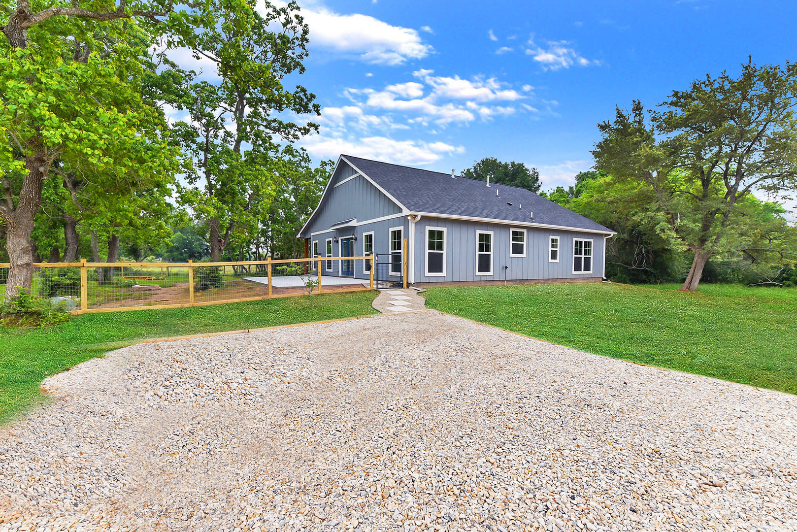 Two-story house with white siding, gravel driveway, wooden fence, mature trees, and grassy lawn under partly cloudy sky