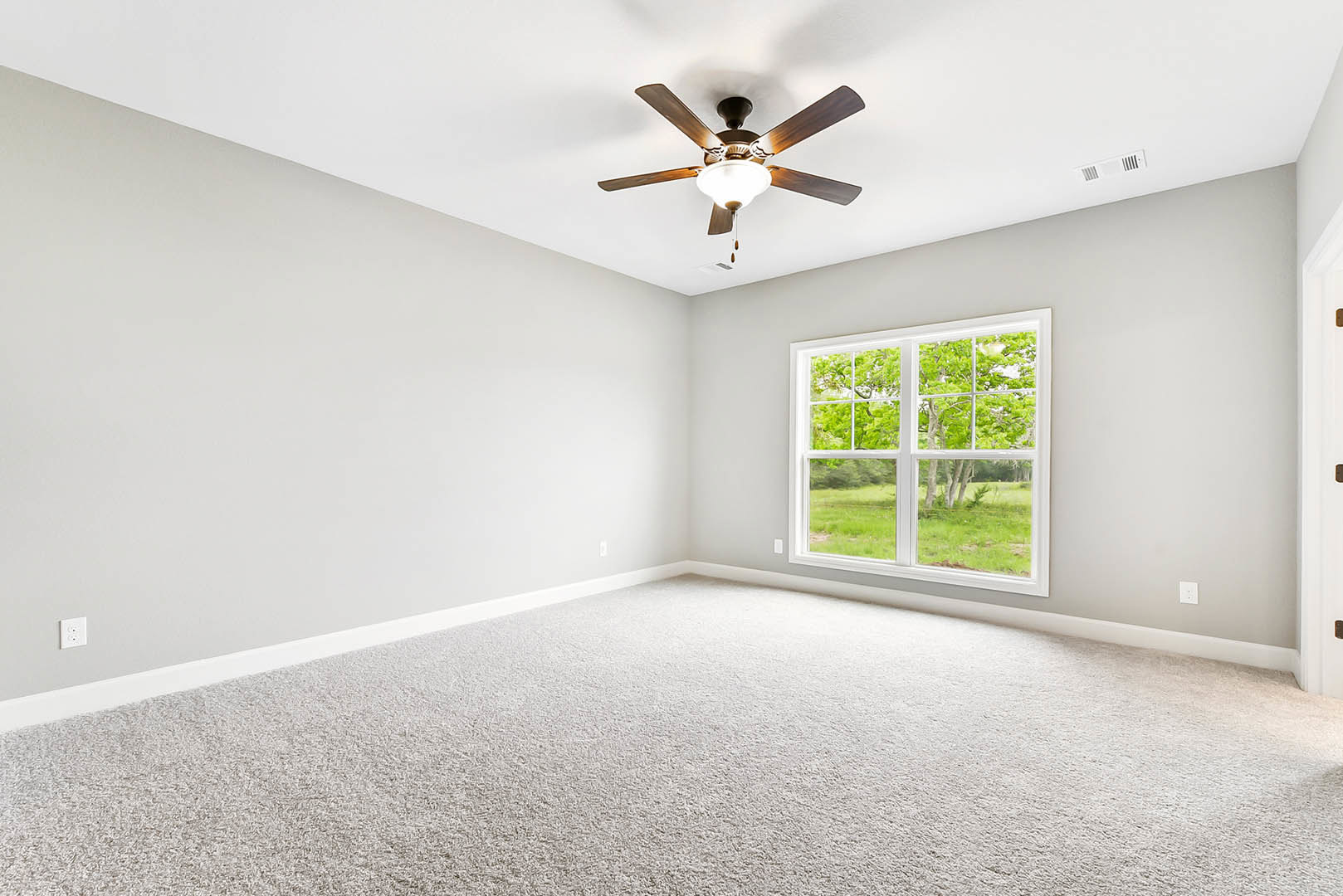 Neutral-toned room with white carpet, ceiling fan with light fixture, large window overlooking green grass and trees, plaster walls and ceiling