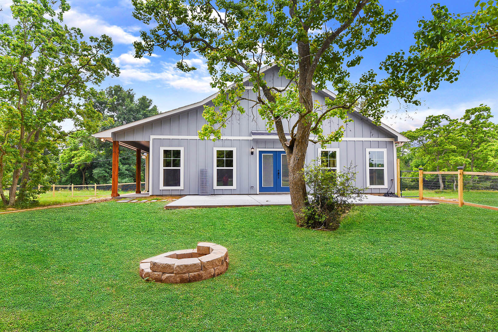 Blue front door with black handle, white-framed window, stone wall and fire pit in grassy backyard, mature tree beside house under partly cloudy sky