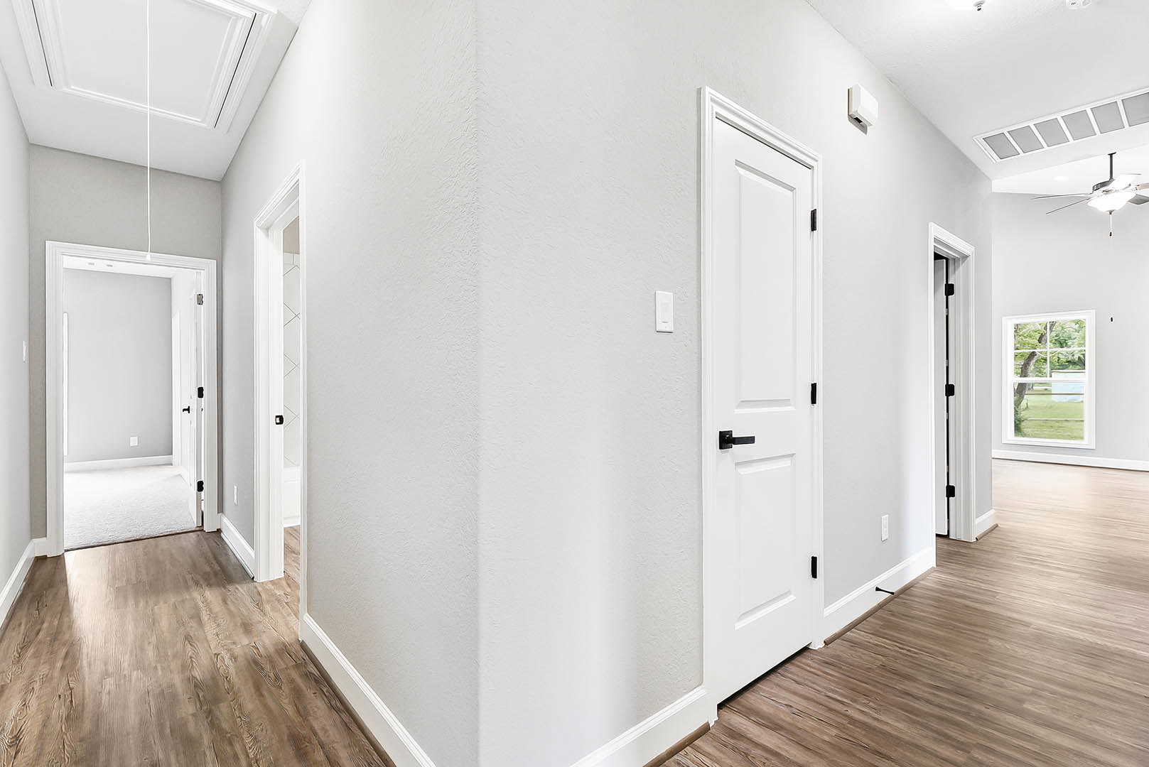 Hallway with wood flooring, white doors featuring black handles, white ceiling, window showing a tree outside, and a white rectangular object with a square hole.