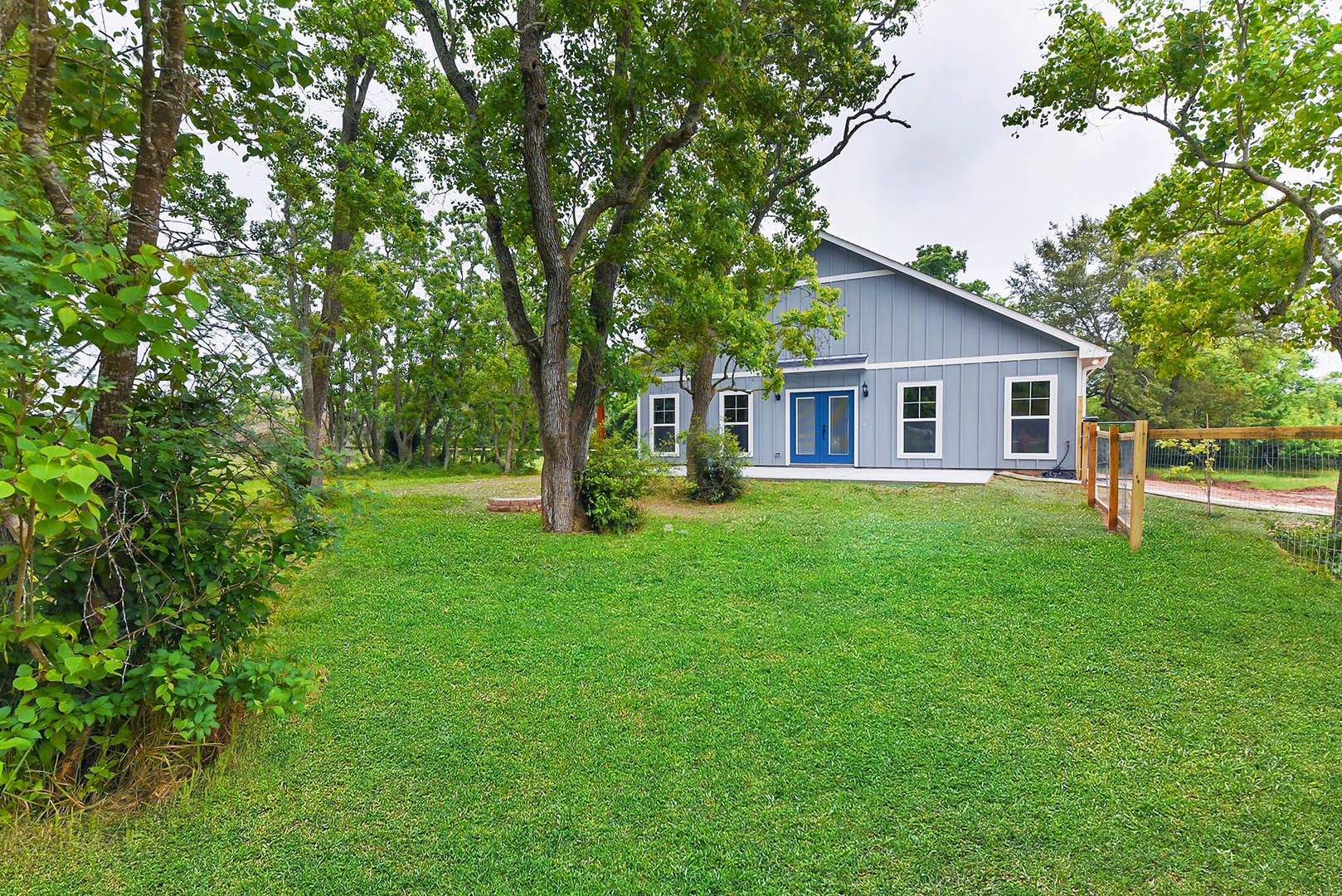 Blue house with white trim, glass-paneled double front door, white-framed windows, fenced yard, green lawn, and mature trees