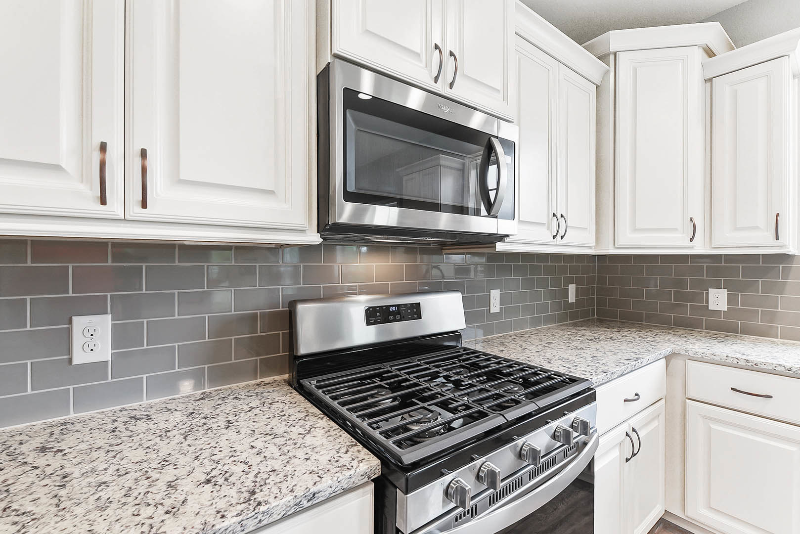 White shaker cabinets, stainless steel gas stove with control knobs, built-in microwave with glass door, quartz countertop, white subway tile backsplash, electrical outlet on wall