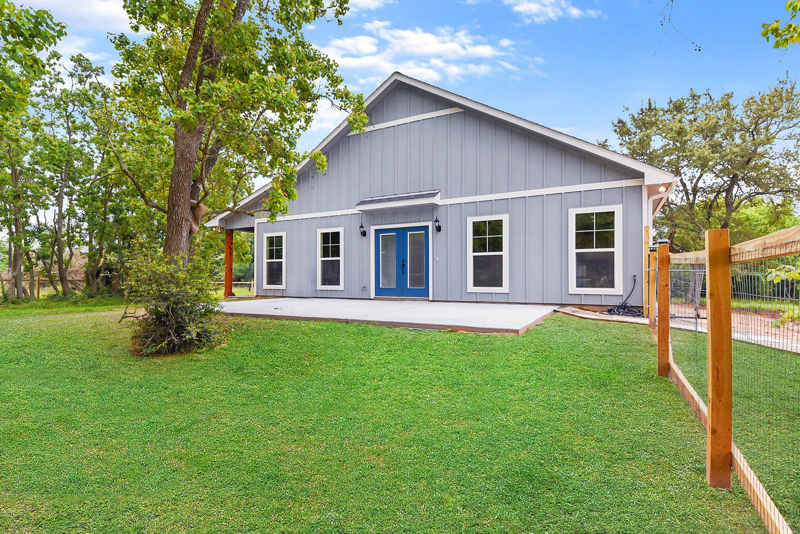 Two-story white house with blue trim and blue glass-paneled front door, large window with white frame, green lawn bordered by sidewalk, mature tree, wooden fence, partly cloudy sky