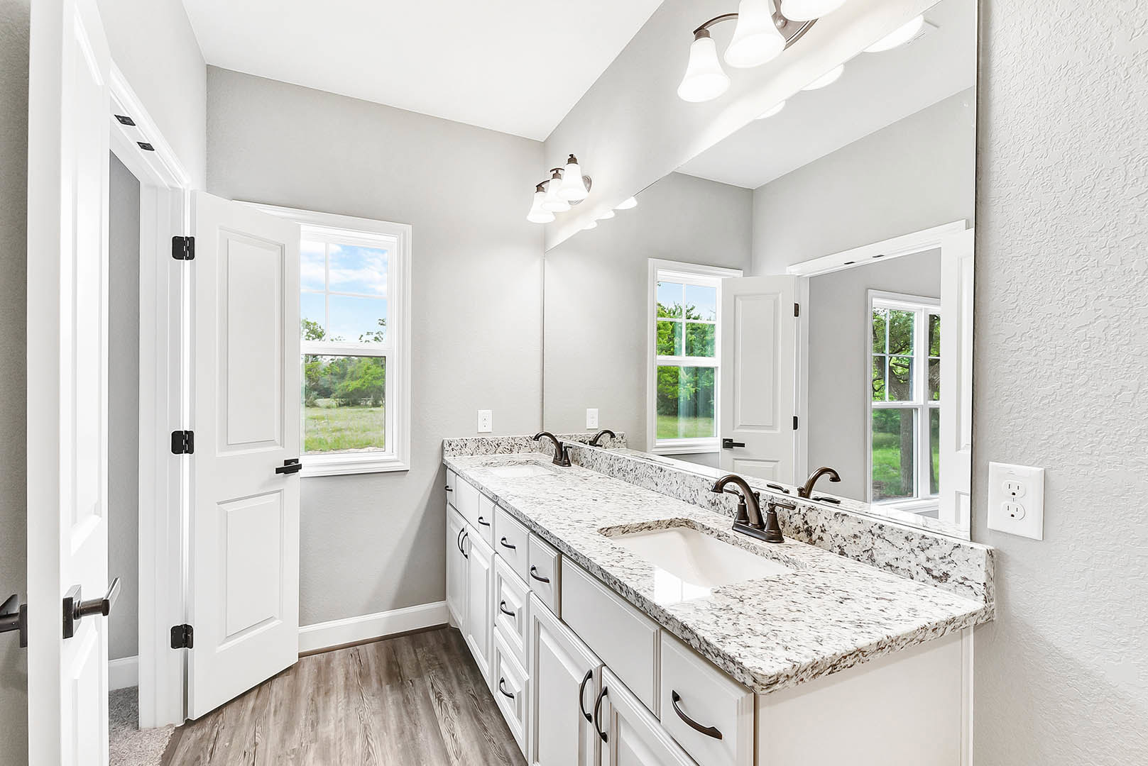 Bathroom with marble countertop, black faucet, double sinks, large mirror, white outlet, white-framed window, and light cabinetry.