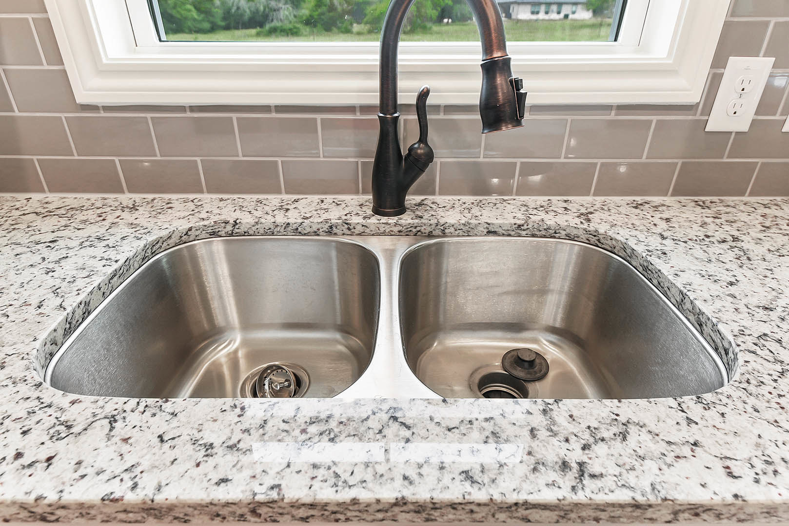 White ceramic sink with chrome faucet set beneath a large window, surrounded by light tile walls and a white electrical outlet.