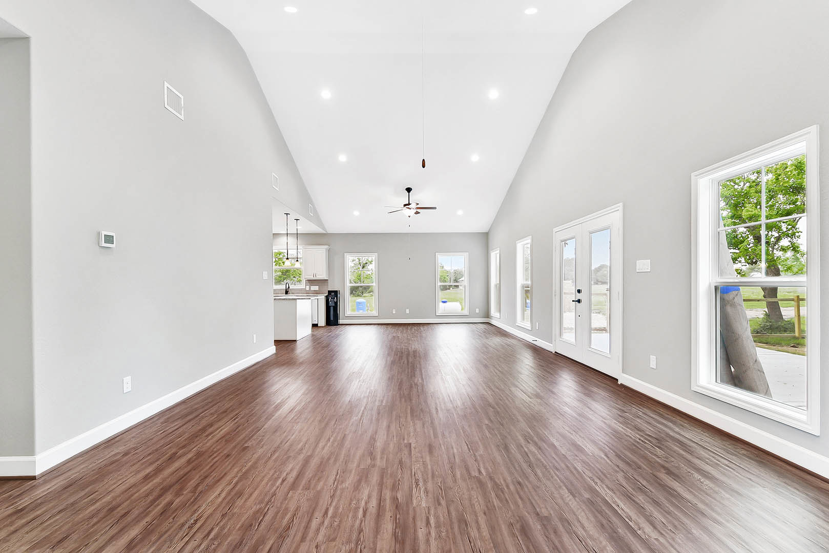 Spacious room with hardwood flooring, white walls, ceiling fan with light, glass-paneled white door, and window overlooking a leafy tree