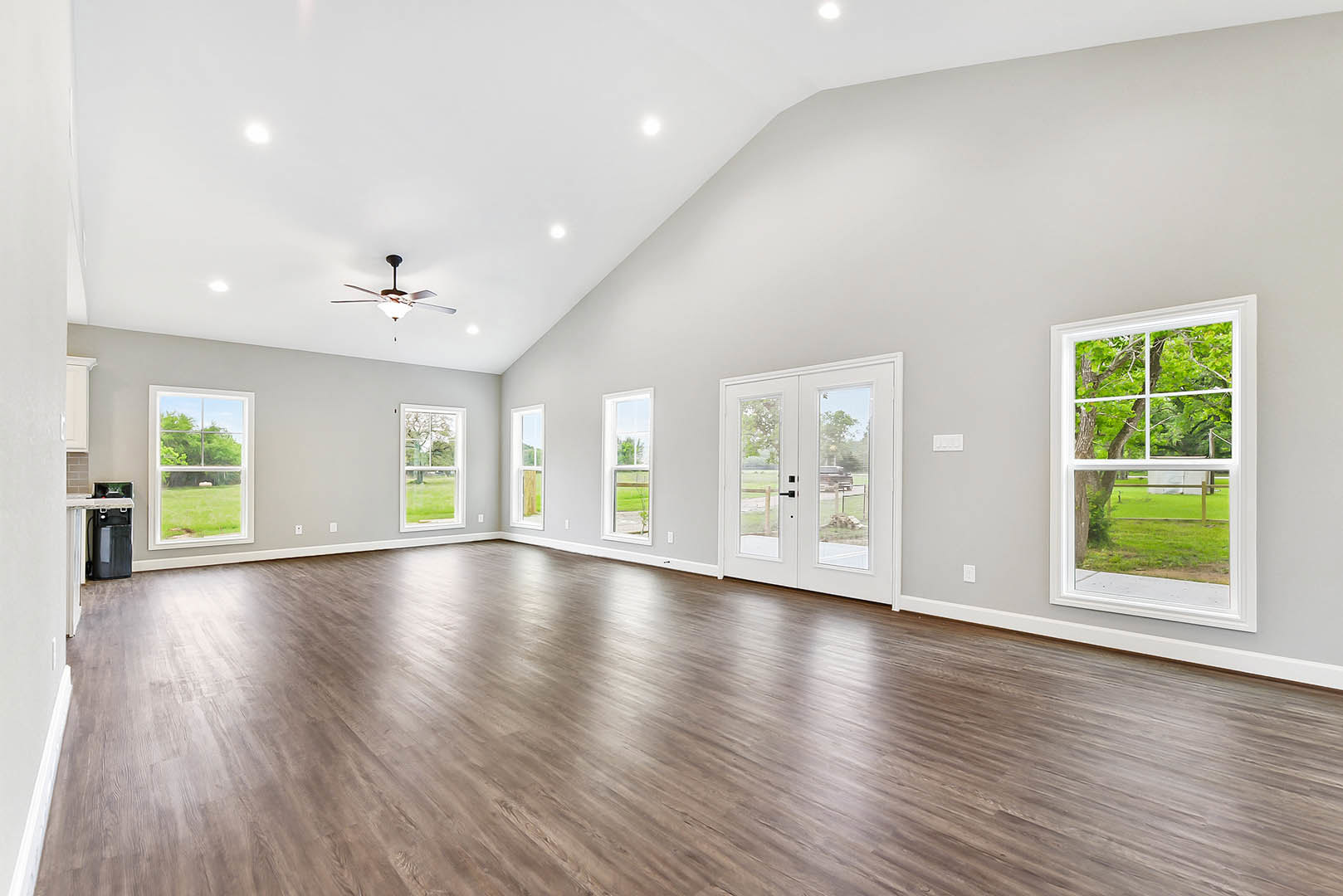 Spacious room with hardwood floors, white double doors with glass panes, ceiling fan with light fixture, large windows overlooking trees