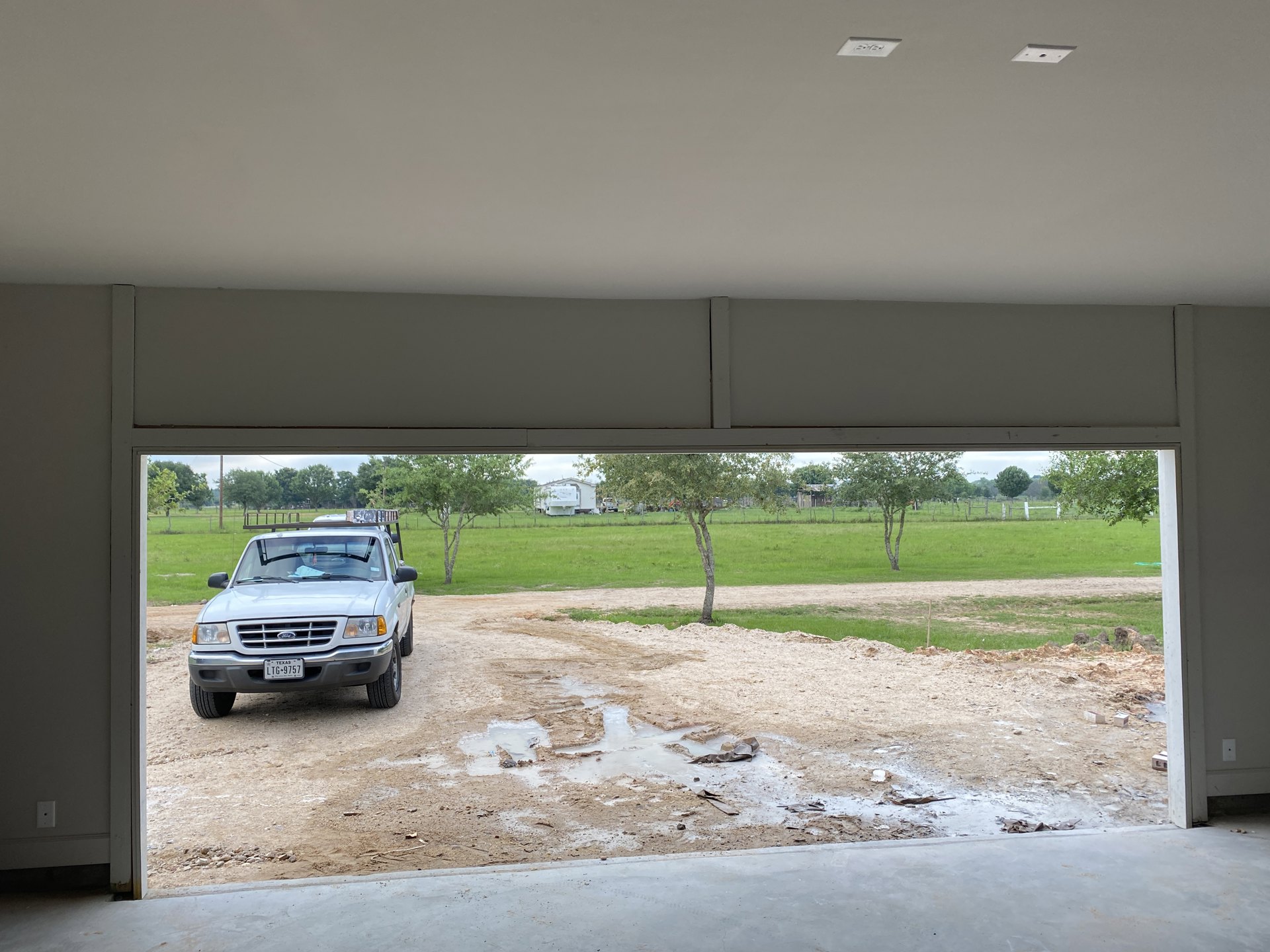 White pickup truck with ladder on roof parked on muddy dirt lot, puddle reflecting tire tracks, grassy patches and grey wall in background