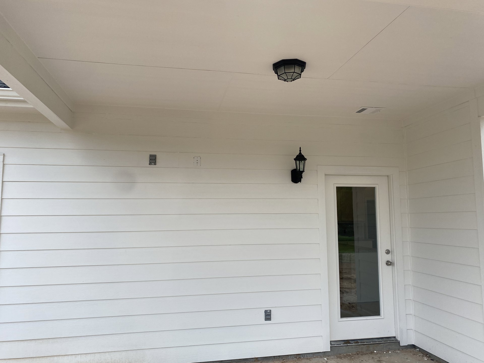 White exterior wall with a black light fixture mounted beside a door featuring a glass panel, beneath a white roof.