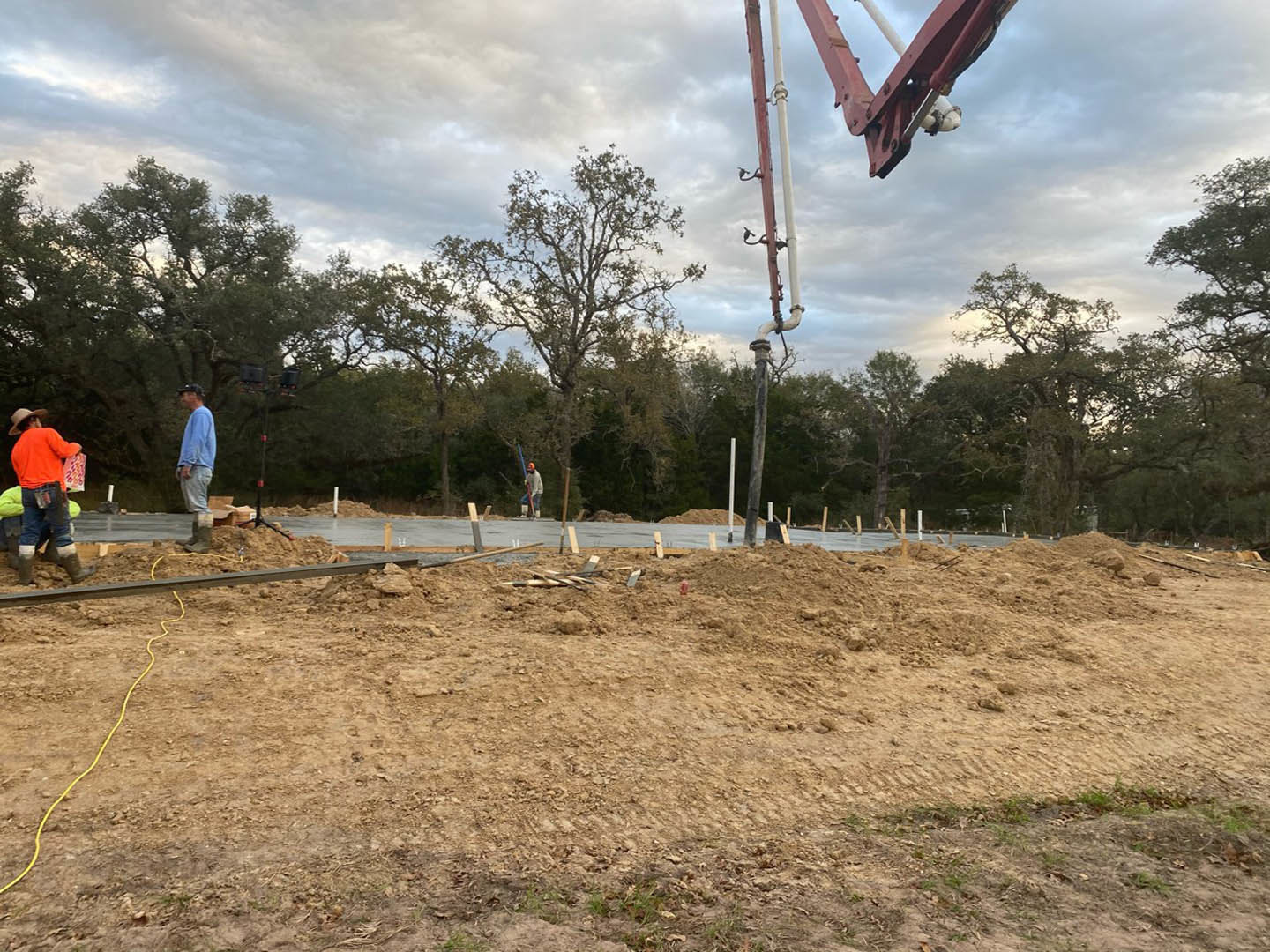 Framed custom home under construction with exposed wood beams, dirt field, crane, scattered lumber, yellow cable, and workers in orange and blue shirts near surrounding trees