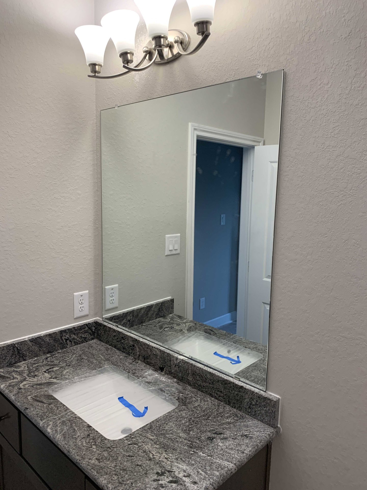 Modern bathroom with white tile walls, rectangular mirror above a quartz countertop, sink covered in plastic wrap and blue tape, three-light fixture, visible outlet and light