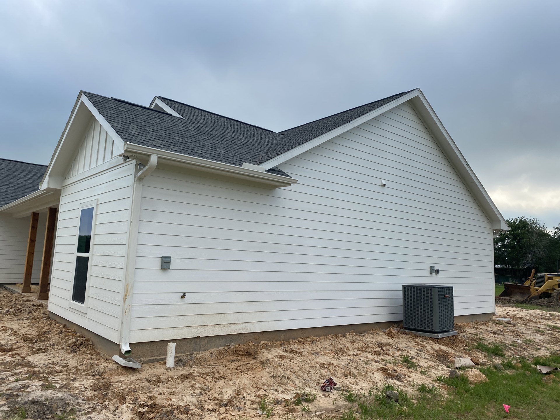 White house with black roof and black box structure on concrete base, large window, dirt landscaping, cloudy sky