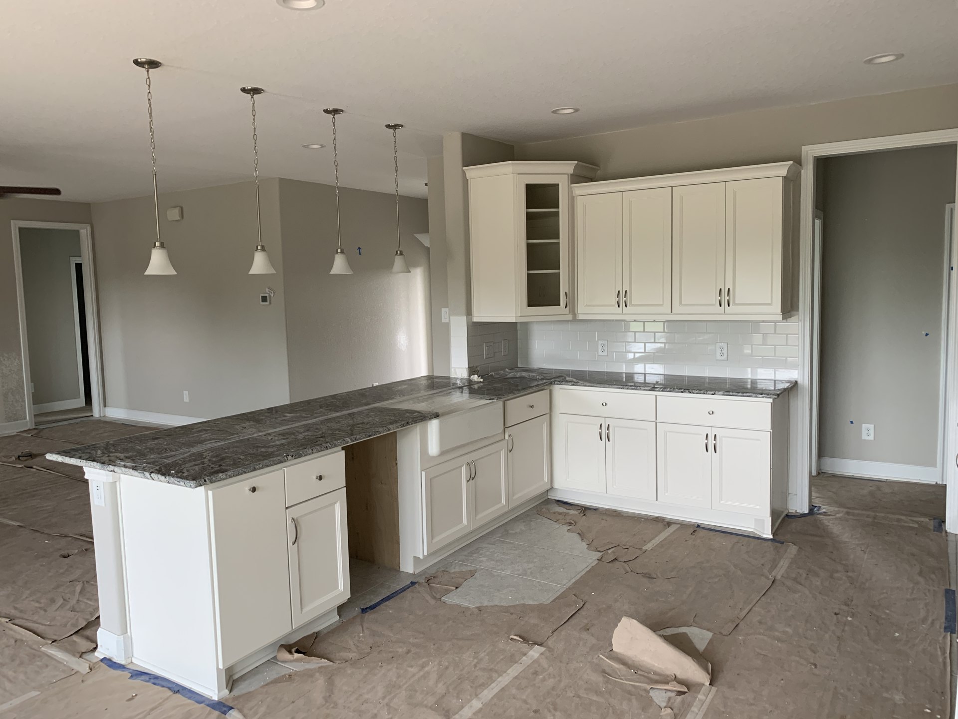 Kitchen with white cabinets featuring silver handles, marble countertops, open shelving, stainless steel sink, white door with black frame, and brown protective paper covering the