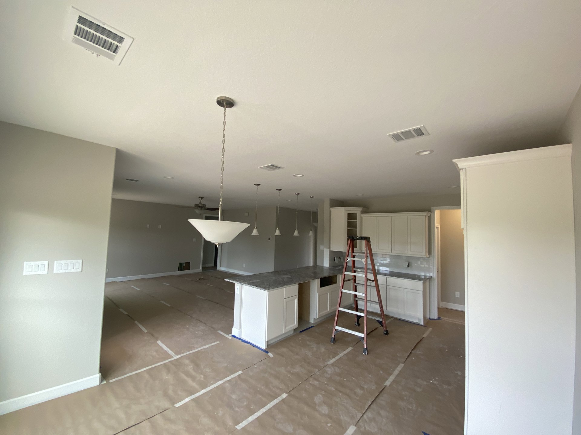 Modern kitchen under construction with a metal ladder positioned on taped floor covering, white plaster walls, ceiling vent, and visible appliance installation.