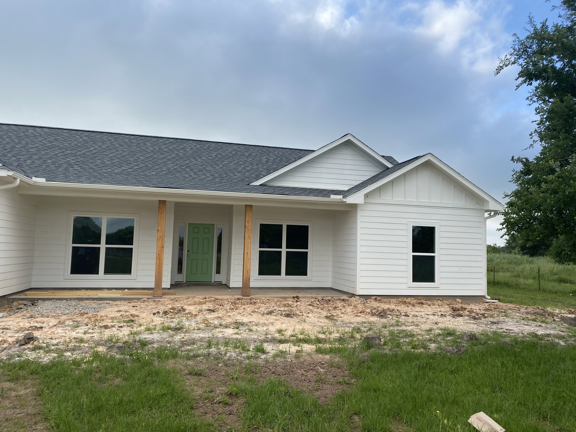White house under construction with green door, white-framed window, wooden pillars, dirt and grass in the yard, cloudy sky overhead