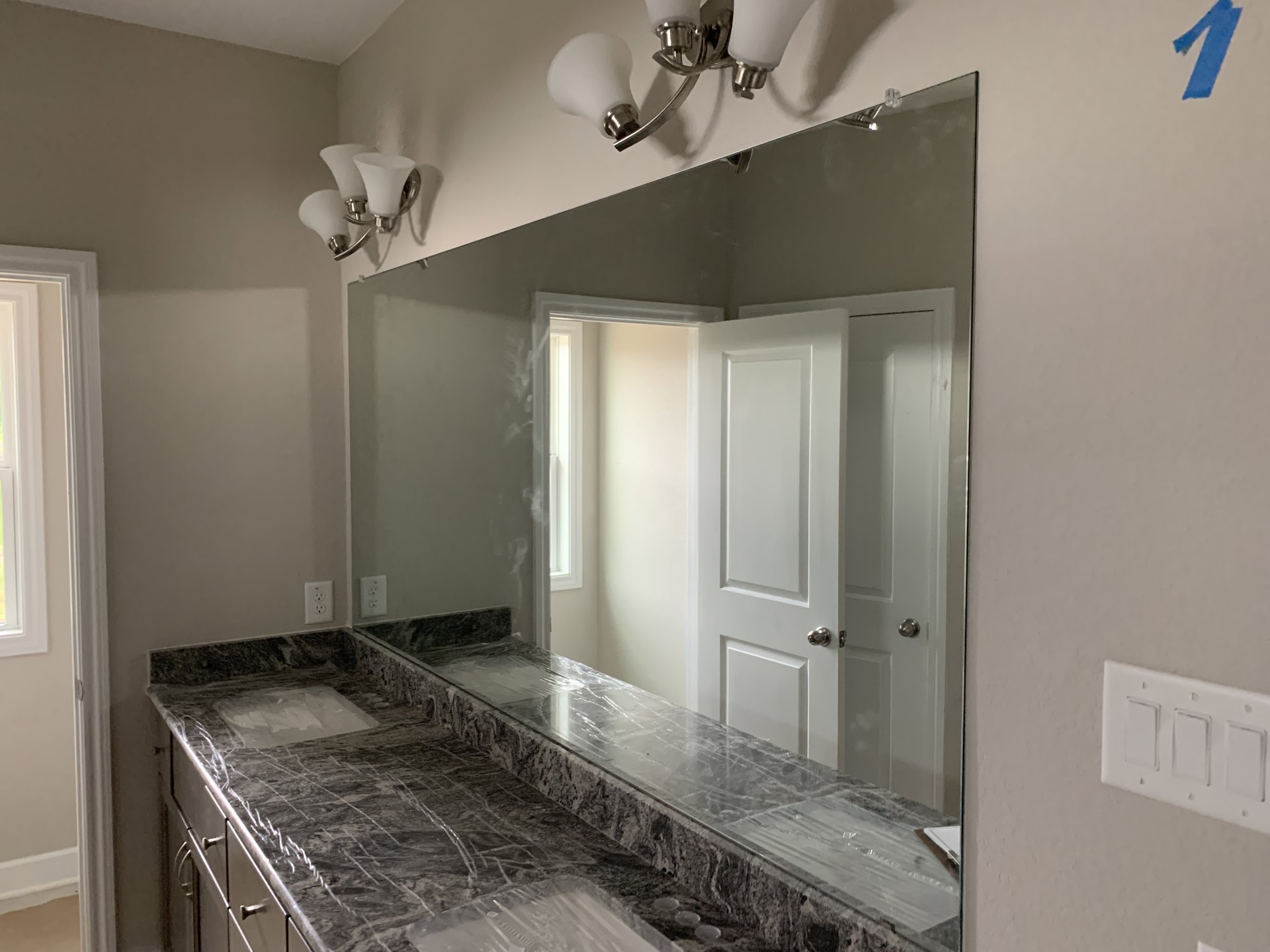 Bathroom with a large wall mirror, white tile walls, light fixture with white glass shades, countertop partially covered in plastic wrap, cabinetry, and a close-up of a light
