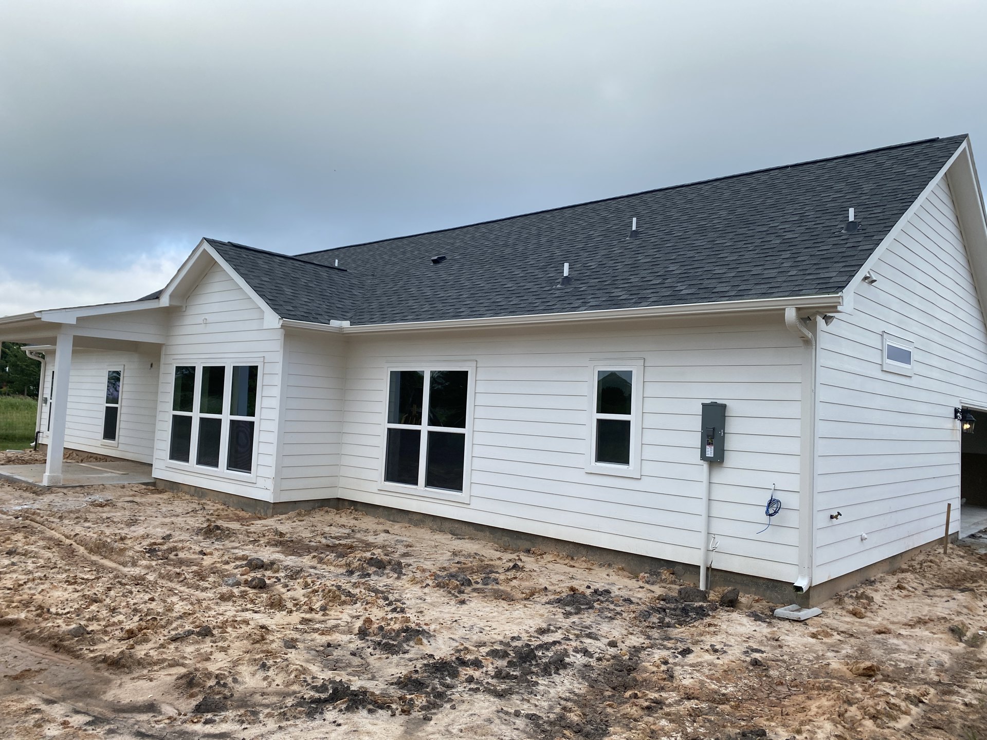 White siding house with black shingle roof, white-framed windows featuring black screens, weather vane mounted atop roof, partly cloudy sky in background