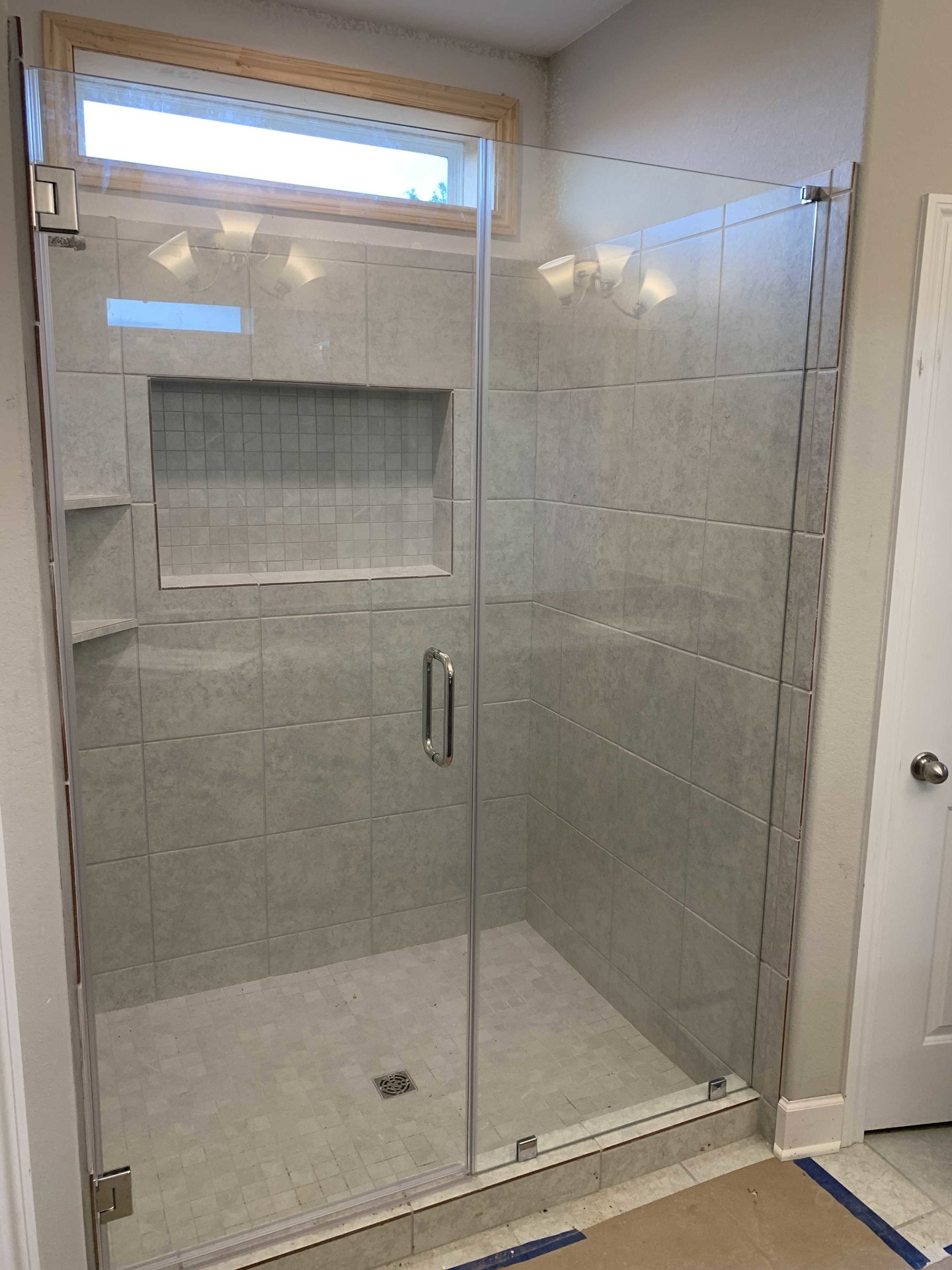 Glass shower enclosure with chrome handle, grey tile walls featuring a rectangular border, window with natural light, and tiled floor in a modern bathroom.