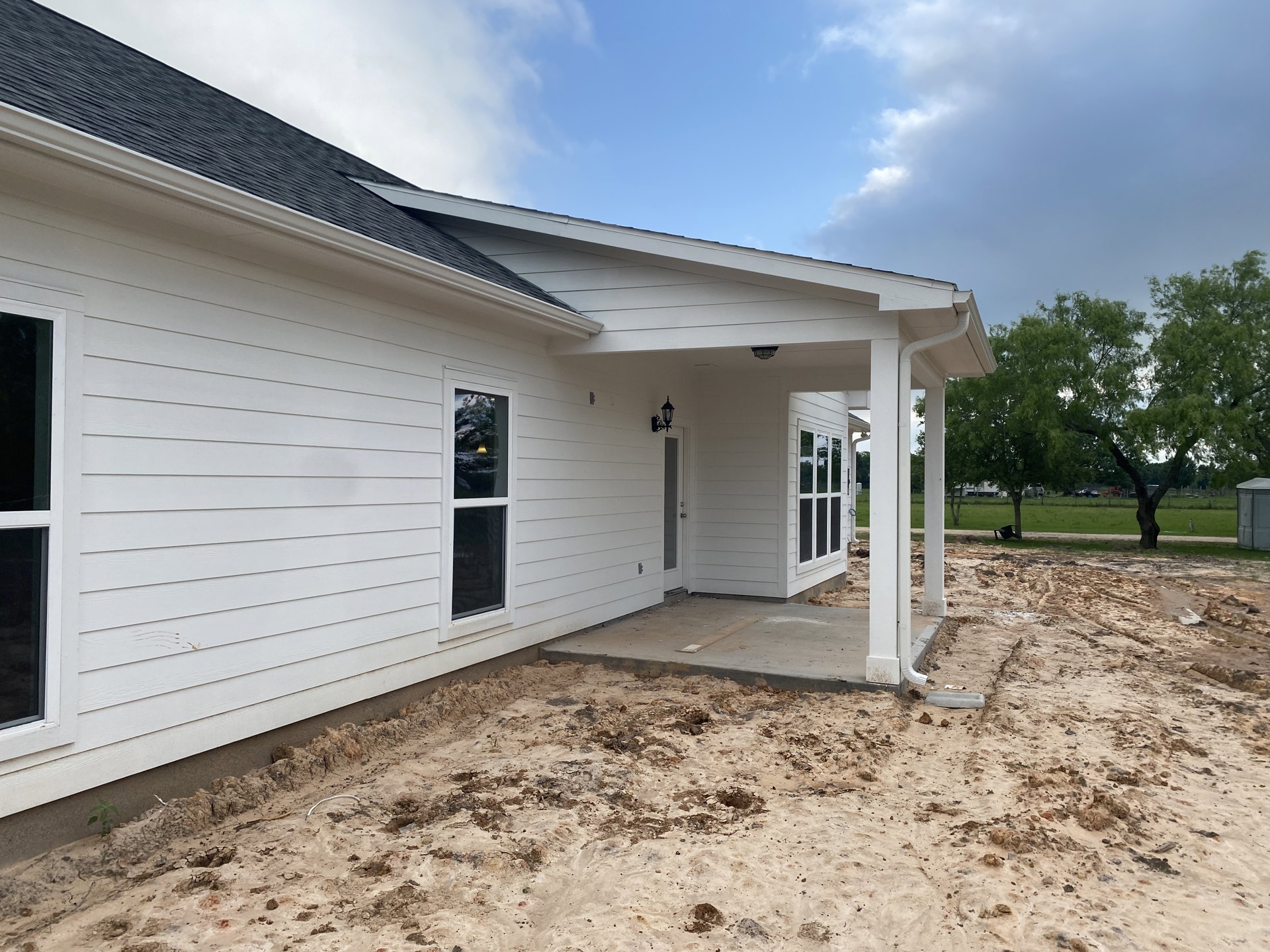 Two-story home with covered porch, large windows reflecting nearby tree, bare dirt yard, illuminated window, cloudy sky overhead