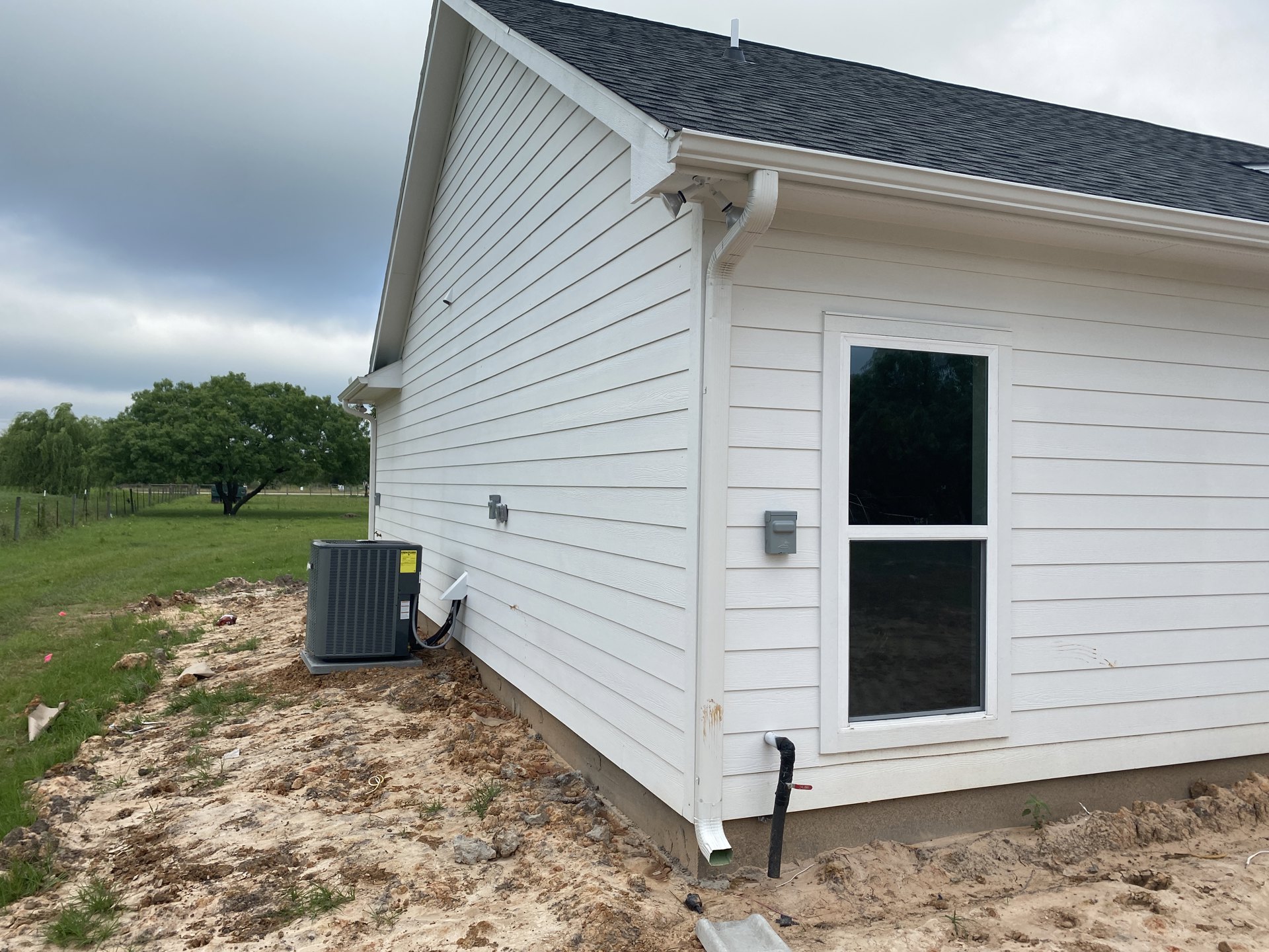 White siding house with black shingle roof, single window framed in black, grassy yard in foreground, cloudy sky above