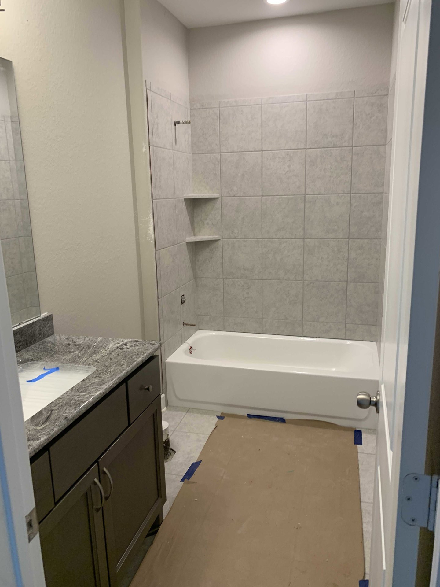 Bathroom with white bathtub taped for construction, cardboard covering floor with blue tape, white tile shower featuring built-in shelf, sink visible, person’s legs raised in