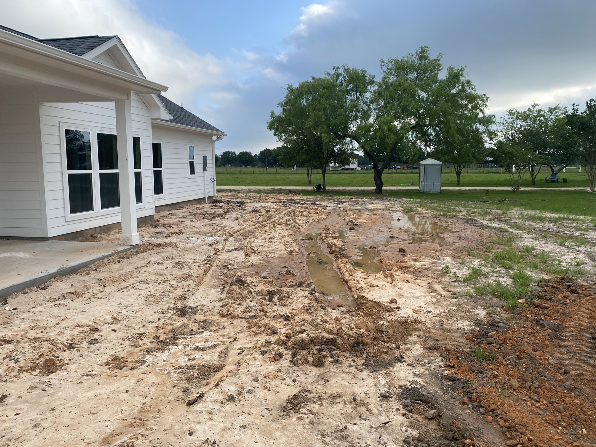 Modern white house with grey roof, white columns, and large windows, surrounded by muddy yard with puddles and sparse grass, tree visible in background under cloudy sky