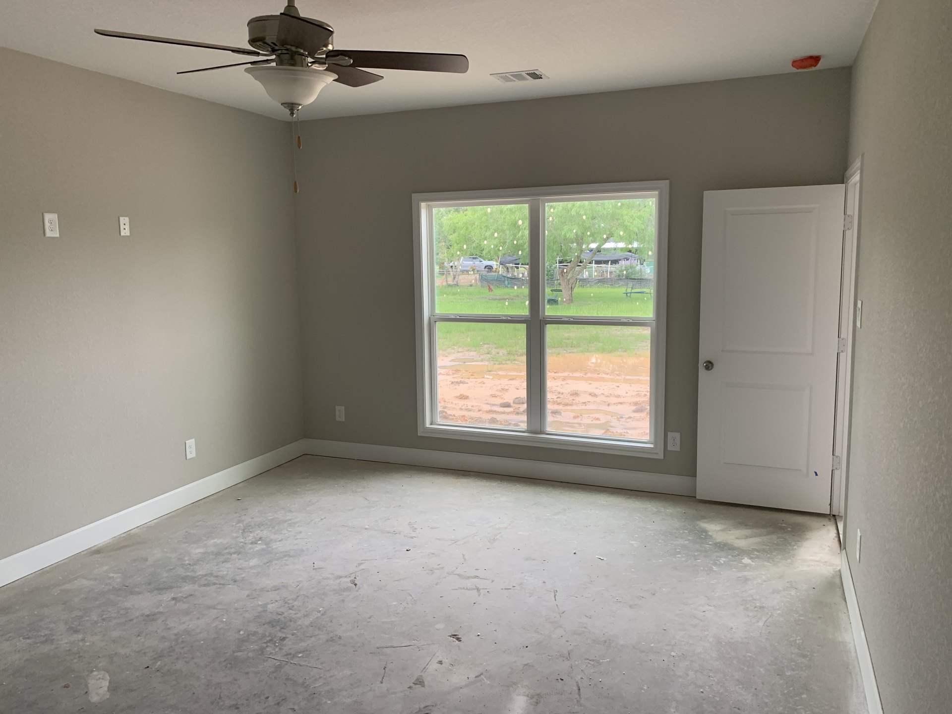Bright room with white walls and floor, ceiling fan with light fixture, large window overlooking yard and trees, white door with silver knob, ceiling vent.